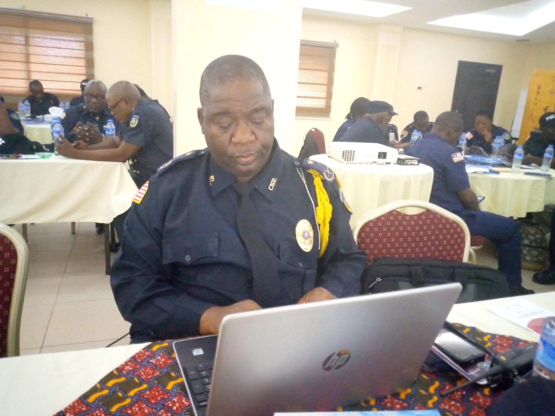 A focused man in a police uniform sits at a laptop in a conference room.