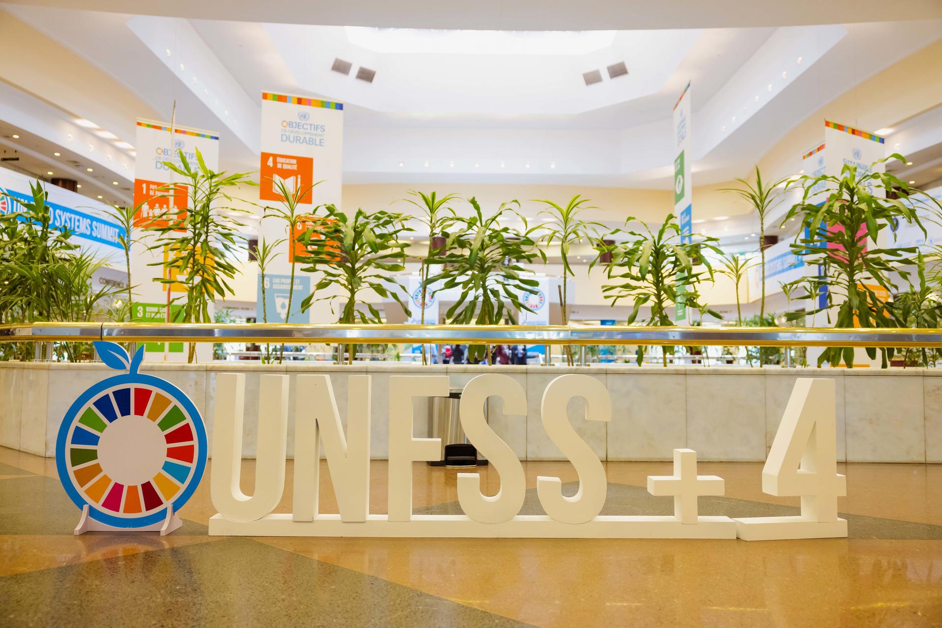 Large white letters spelling "UNESS ± 4" with a colorful logo, surrounded by indoor plants.