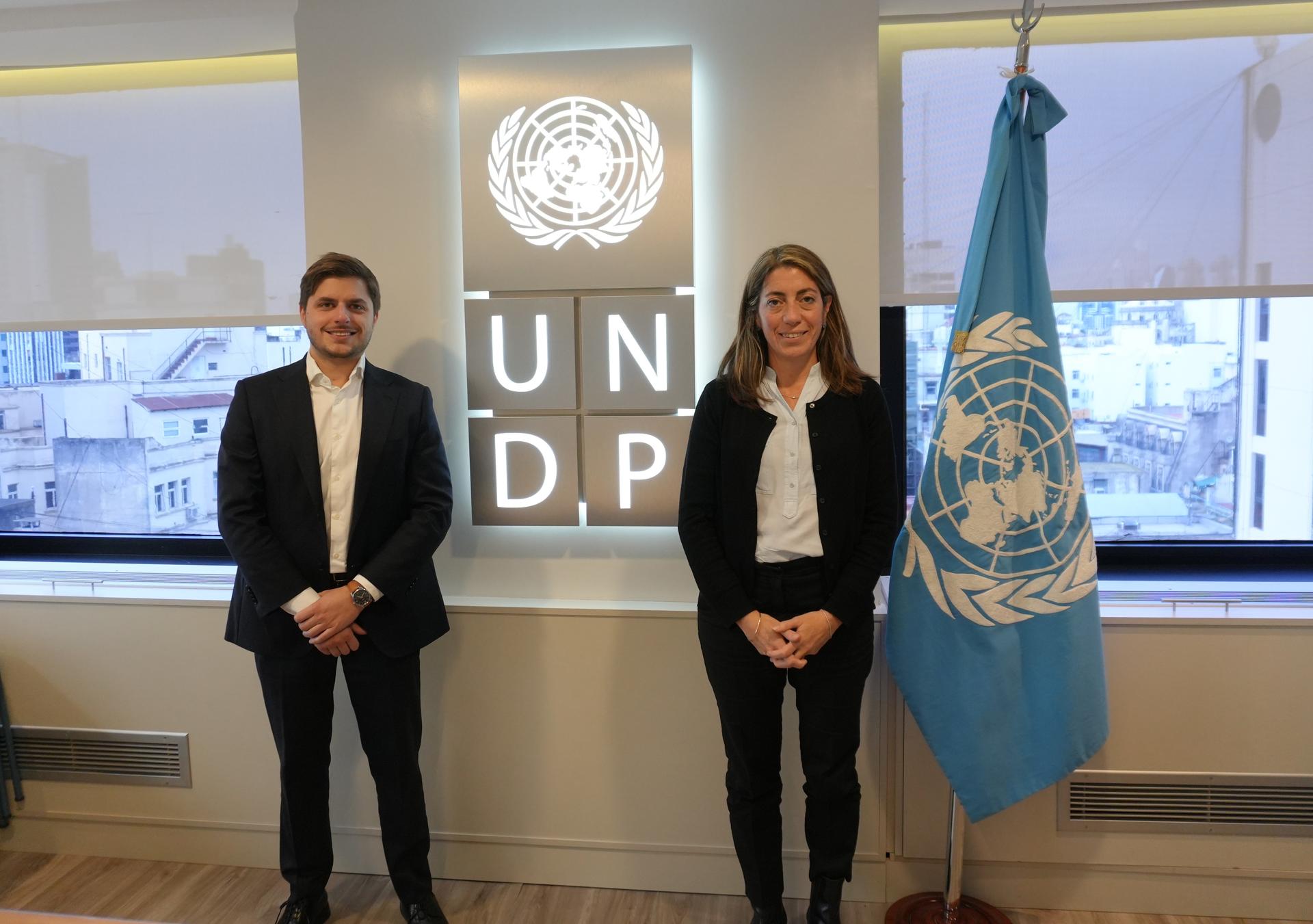 Un hombre y una mujer vestidos formalmente parados frente a un logo de PNUD y junto a una bandera de Naciones Unidas en un entorno de oficina..