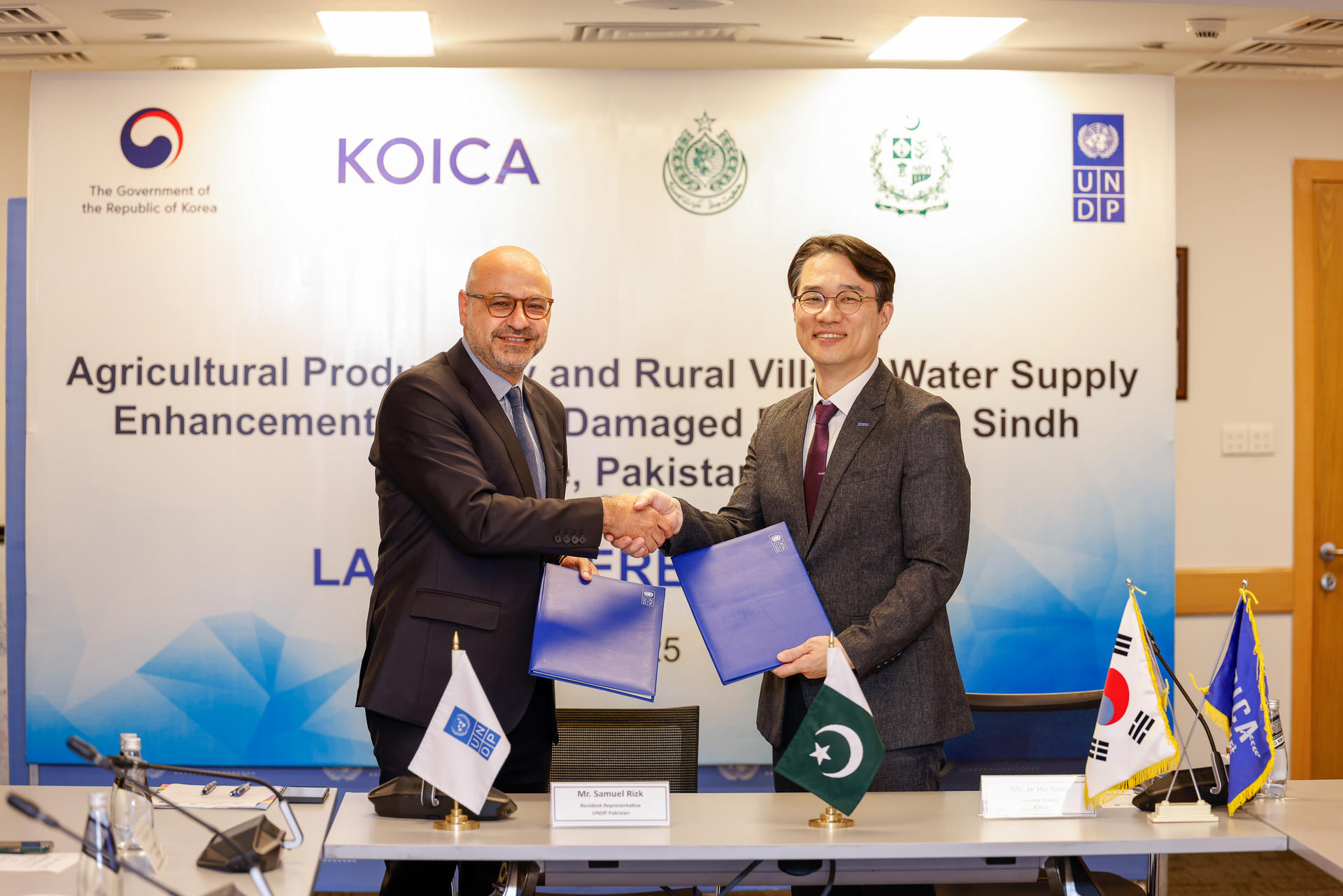 Two men shake hands while holding signed documents, with a backdrop featuring logos and text.