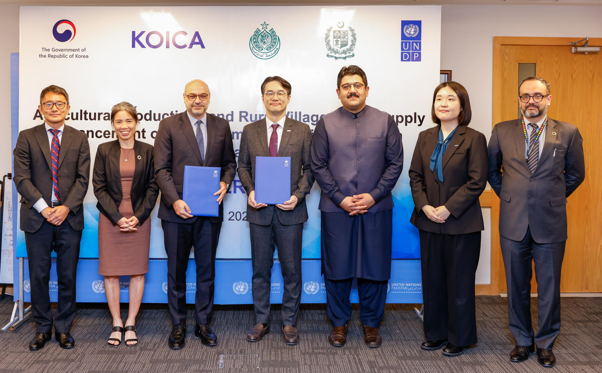 Group photo of eight people in formal attire, holding documents, in an office setting.