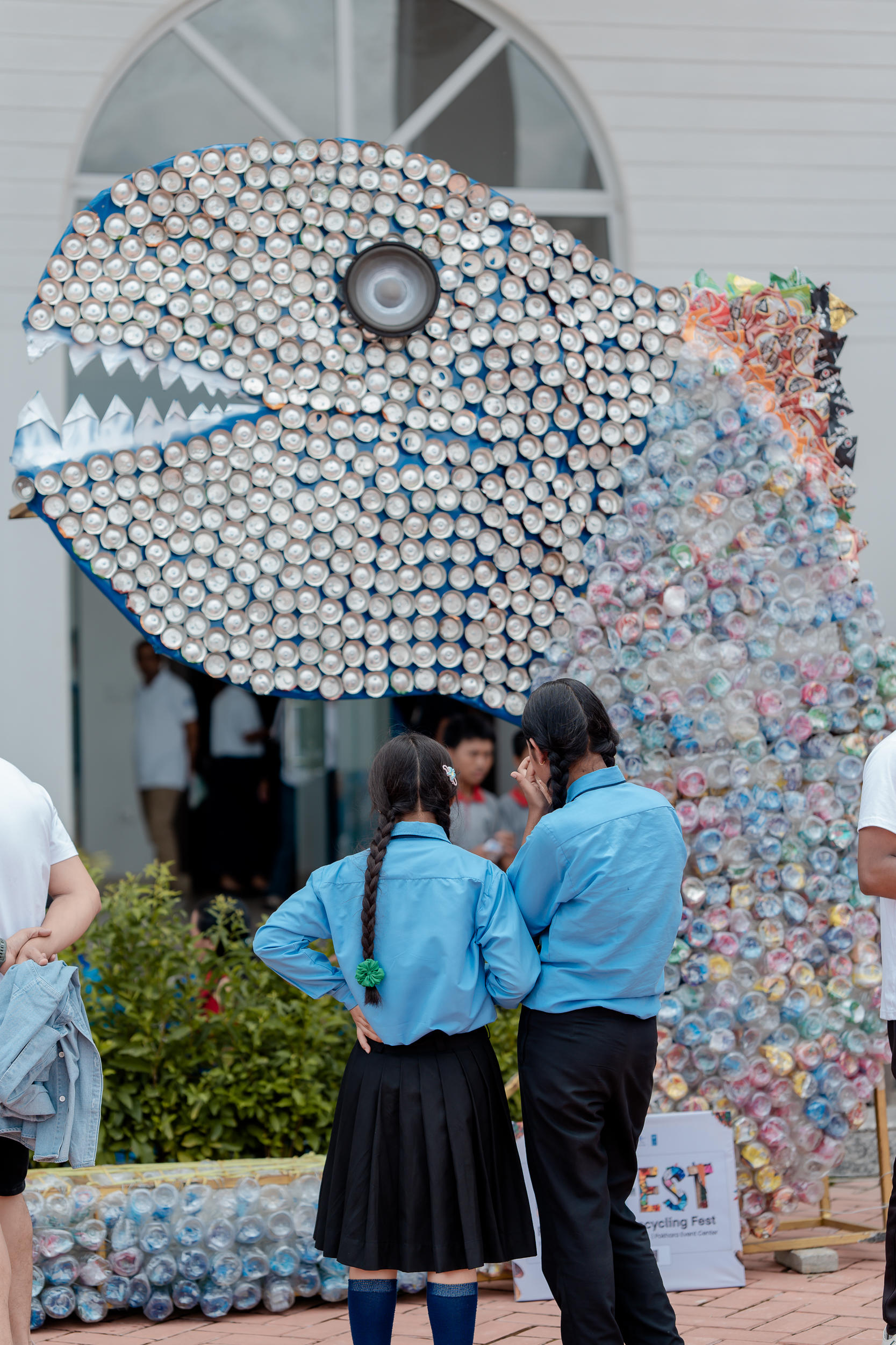 Two people in blue shirts observe a large sculpture of a fish made from cans.
