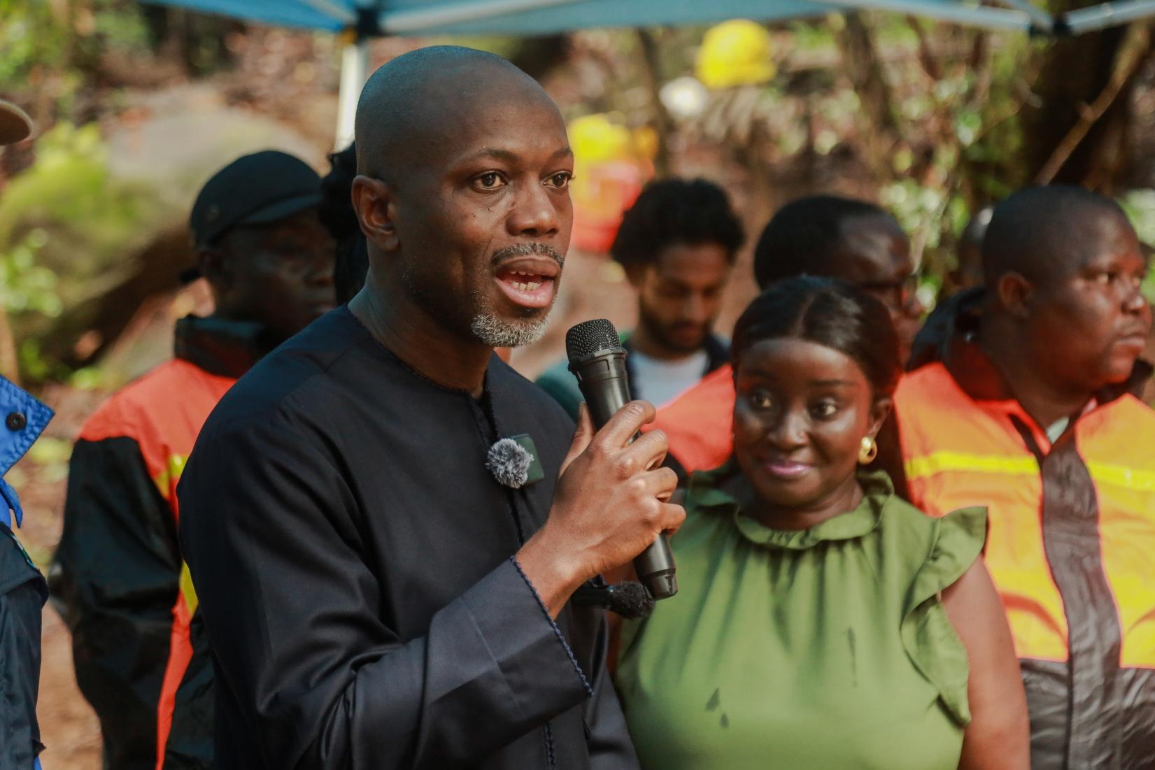 A man speaks into a microphone, with a woman smiling beside him and others in the background.