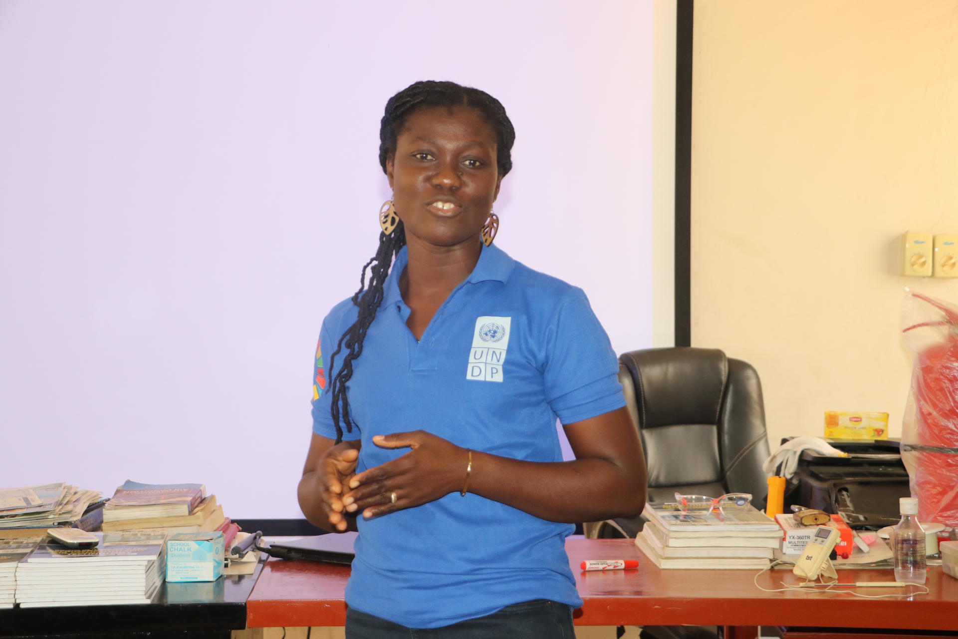 A woman in a blue shirt speaks to an audience in a classroom setting.