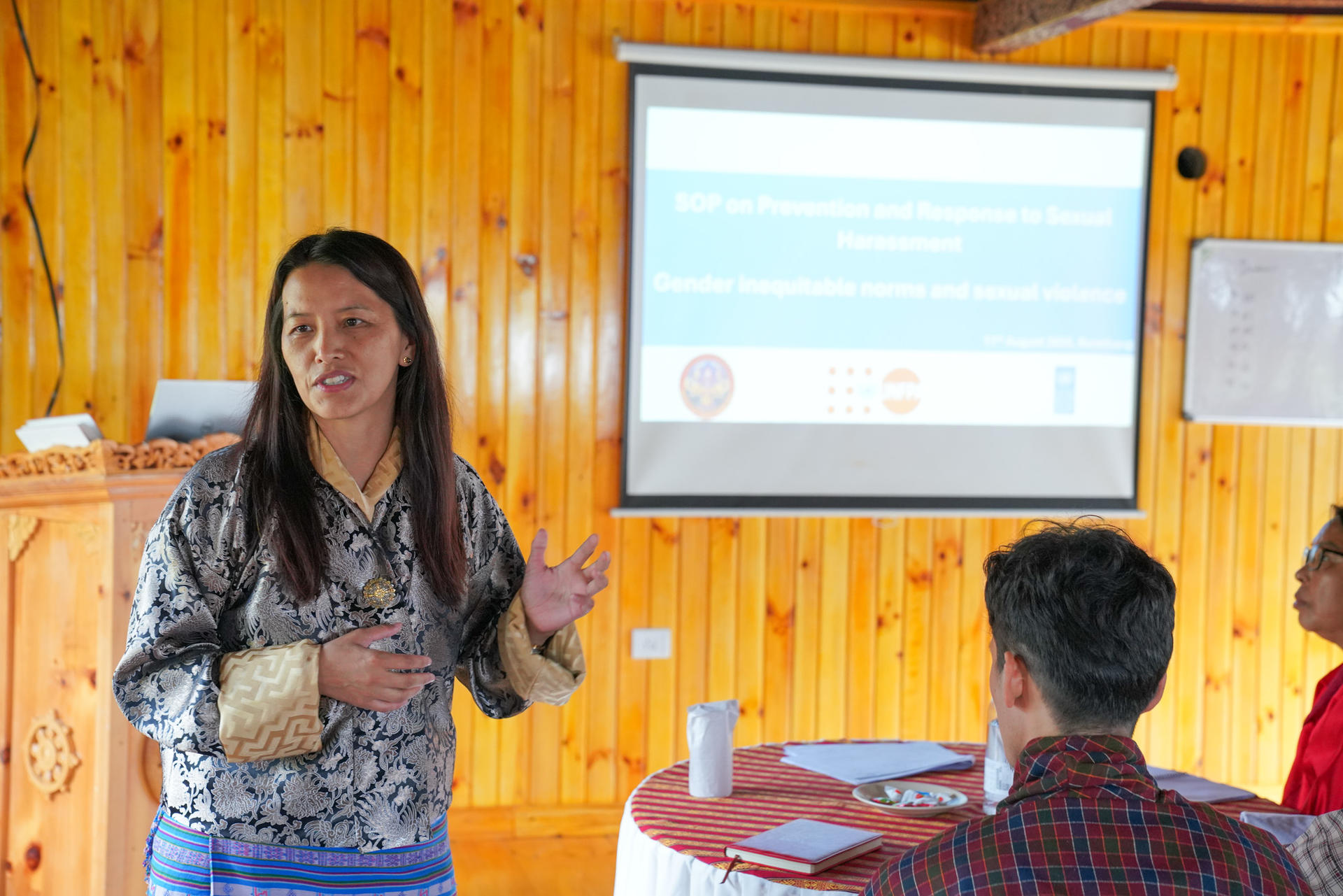 A woman in traditional attire presents to an audience in a wooden room.