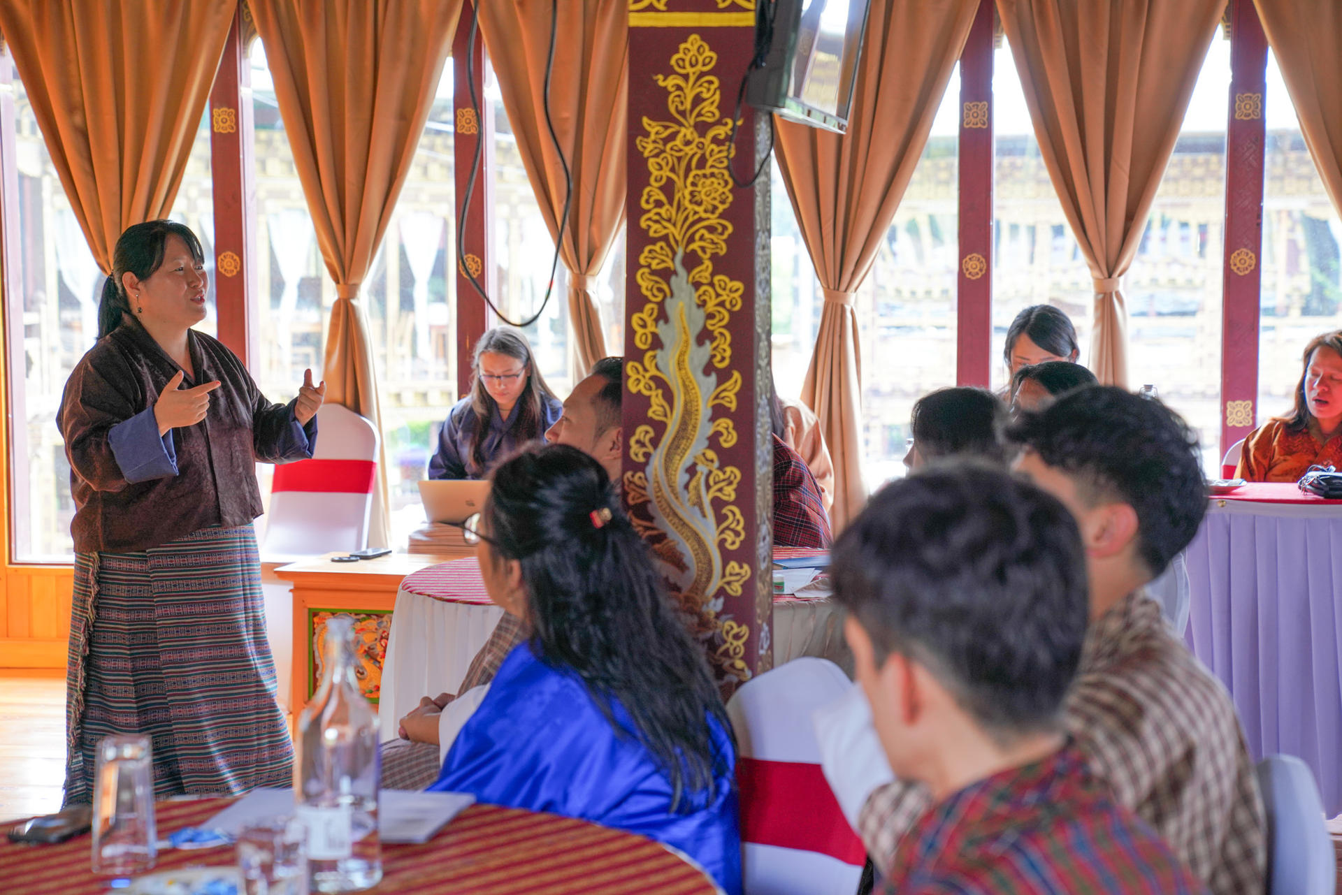 A speaker gestures while presenting to an engaged audience in a decorated hall.