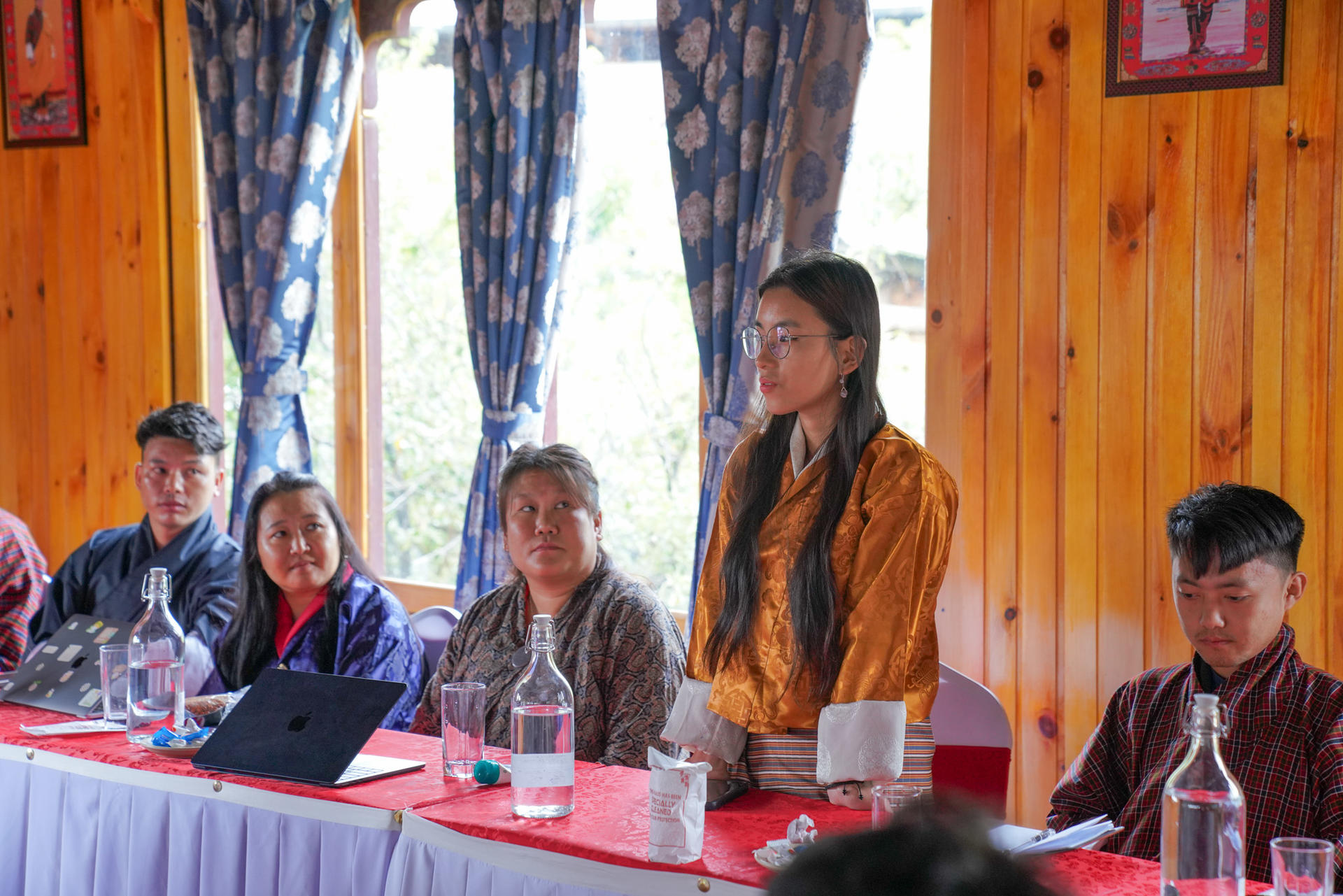 A woman in an orange garment stands at a table, speaking to a group in a wooden room.