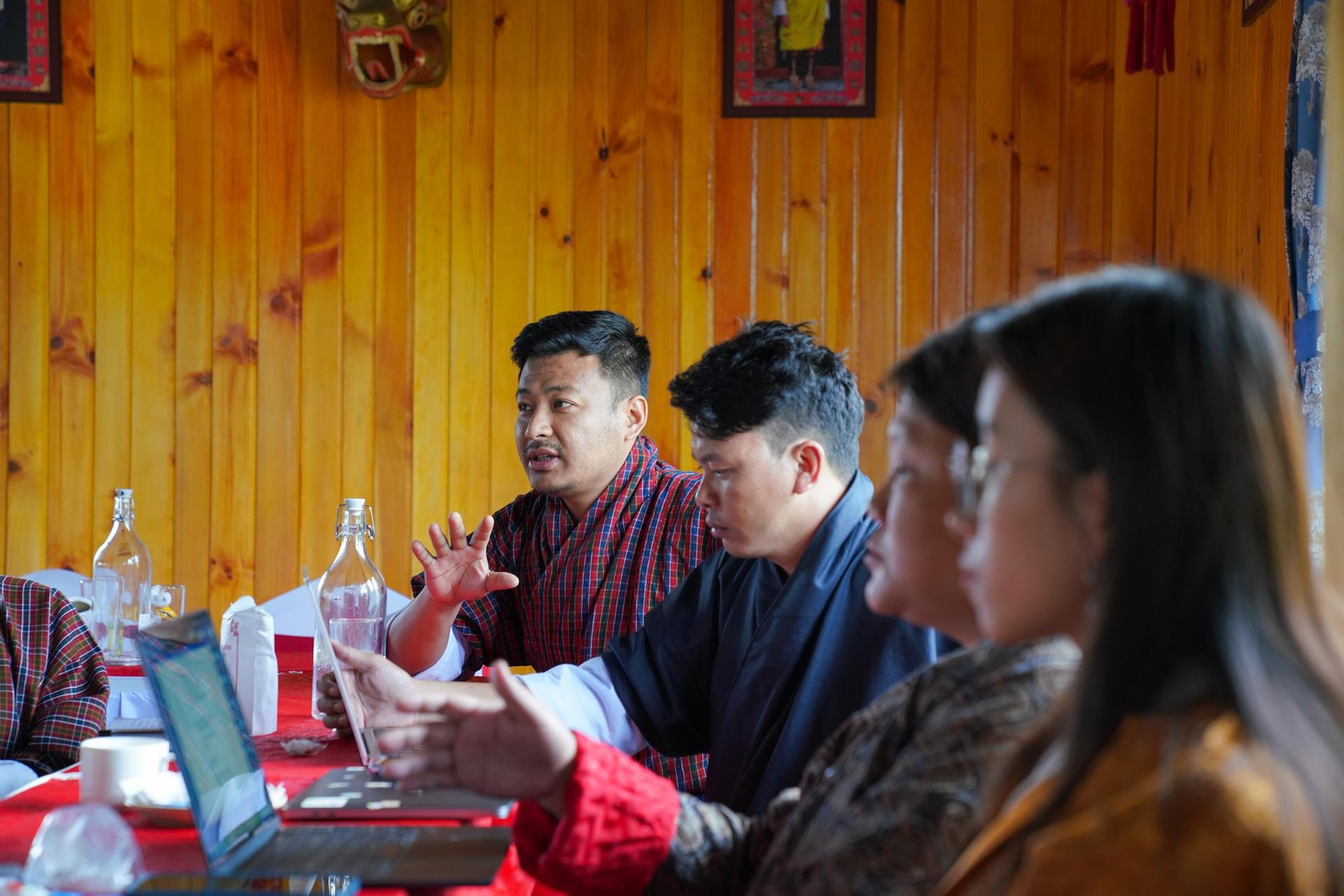 A group of four people engaged in discussion at a conference table in a wooden-paneled room.