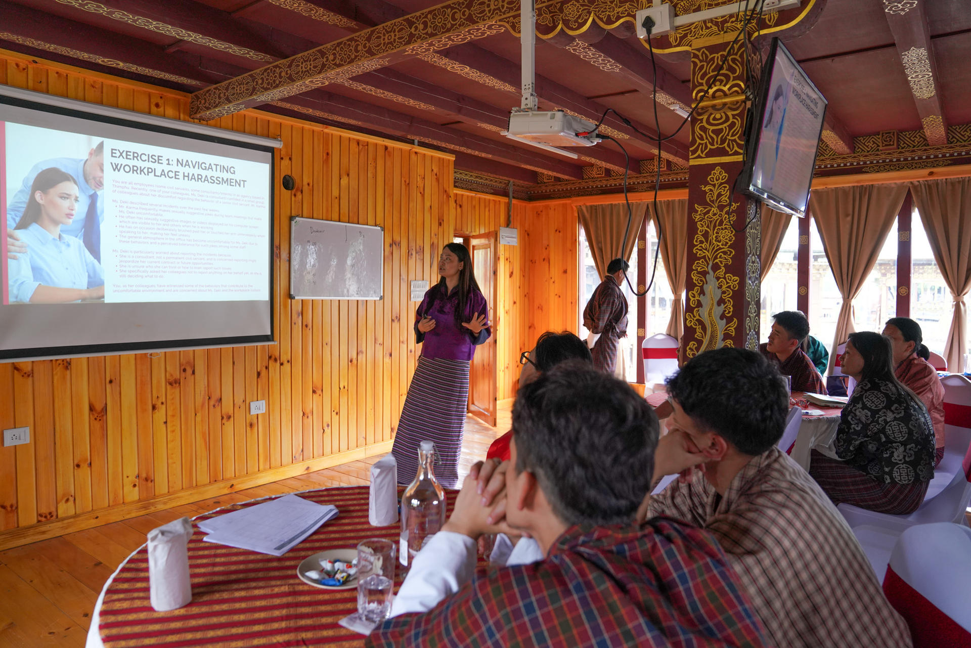 A woman presents to an audience in a wooden conference room, with visible screens and seating.