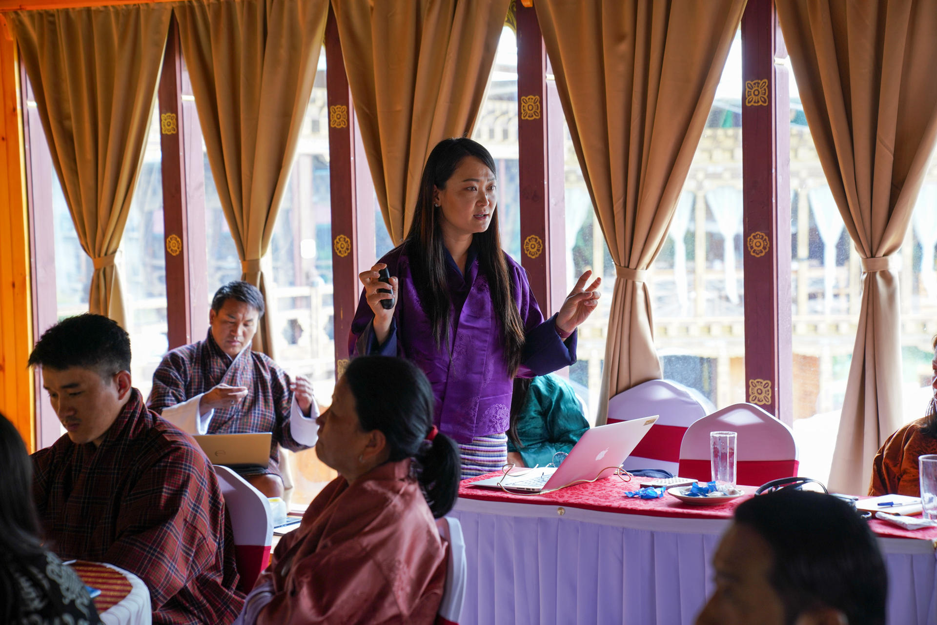 A woman in traditional attire gestures while speaking to an audience in a brightly lit room.