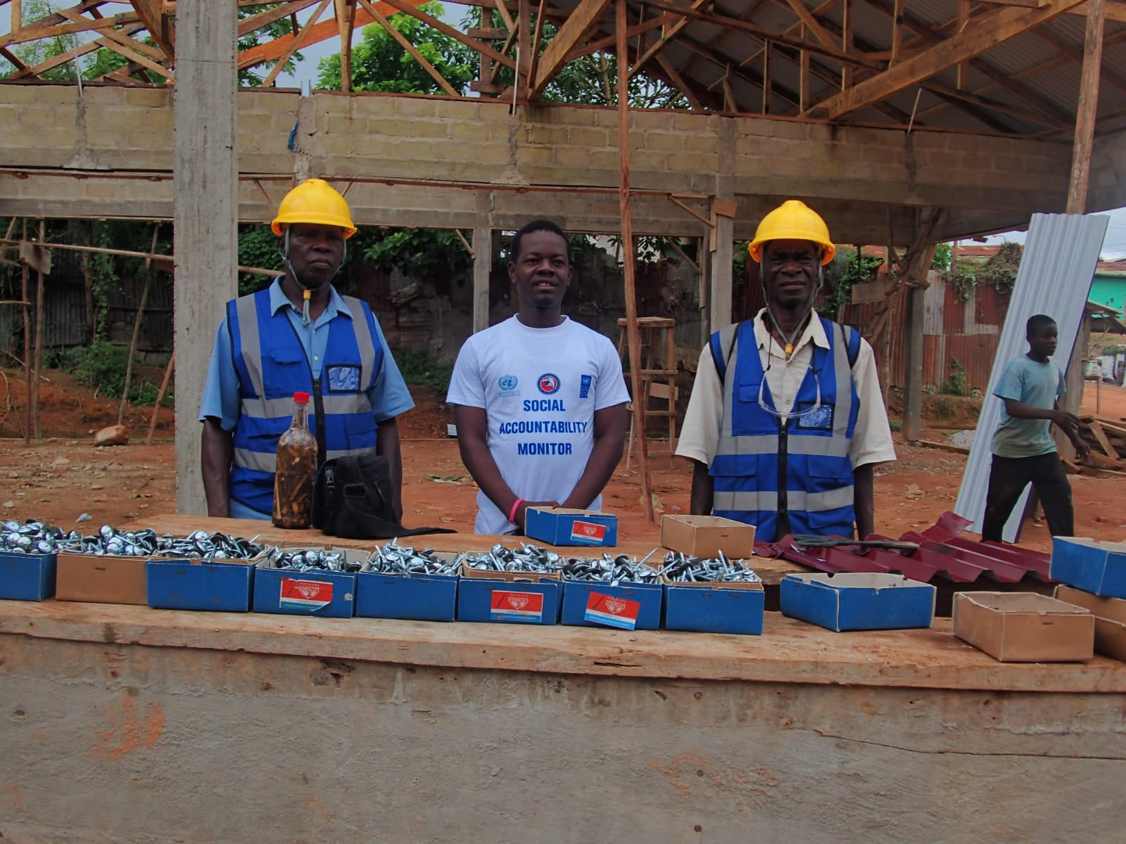 Three men in hard hats and vests stand at a construction site with tools and materials.
