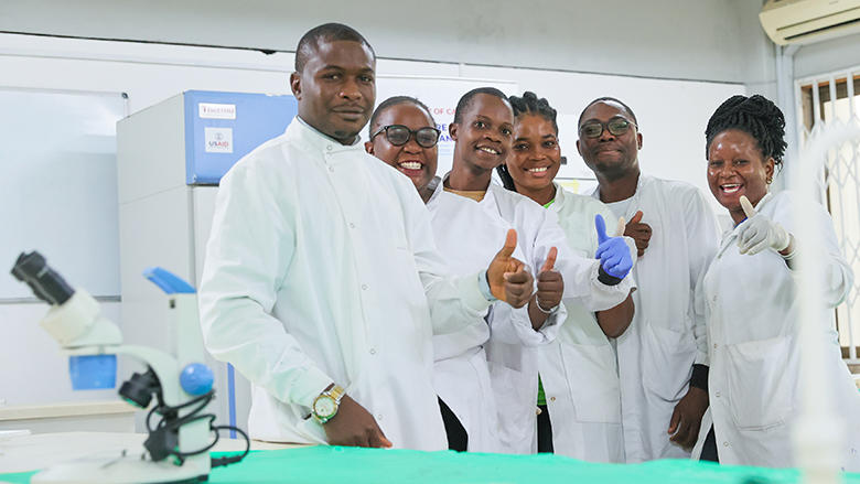 A group of six people in lab coats smiling and giving thumbs up in a laboratory setting.