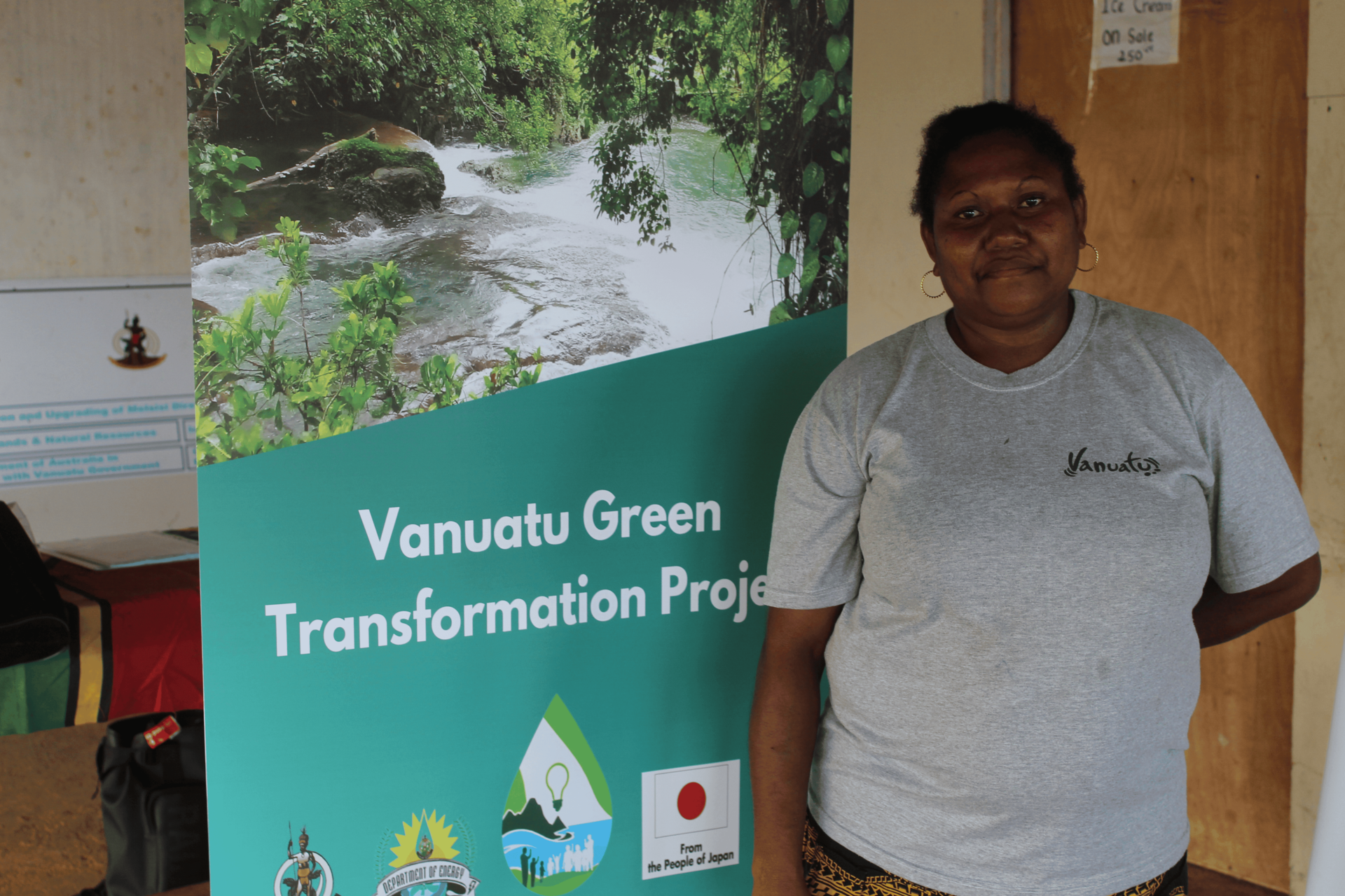 A woman stands beside a banner featuring a river and greenery for the Vanuatu Green Transformation Project.