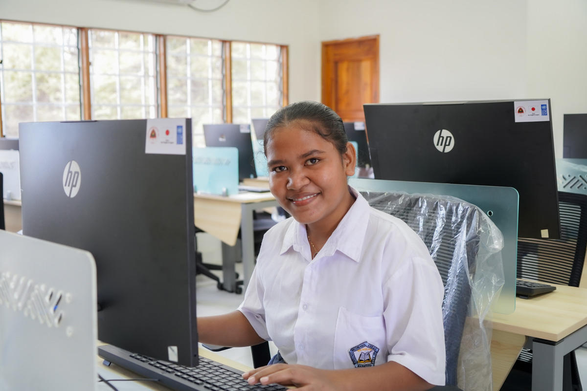 A smiling student in a school computer lab, sitting at a desk with a computer.