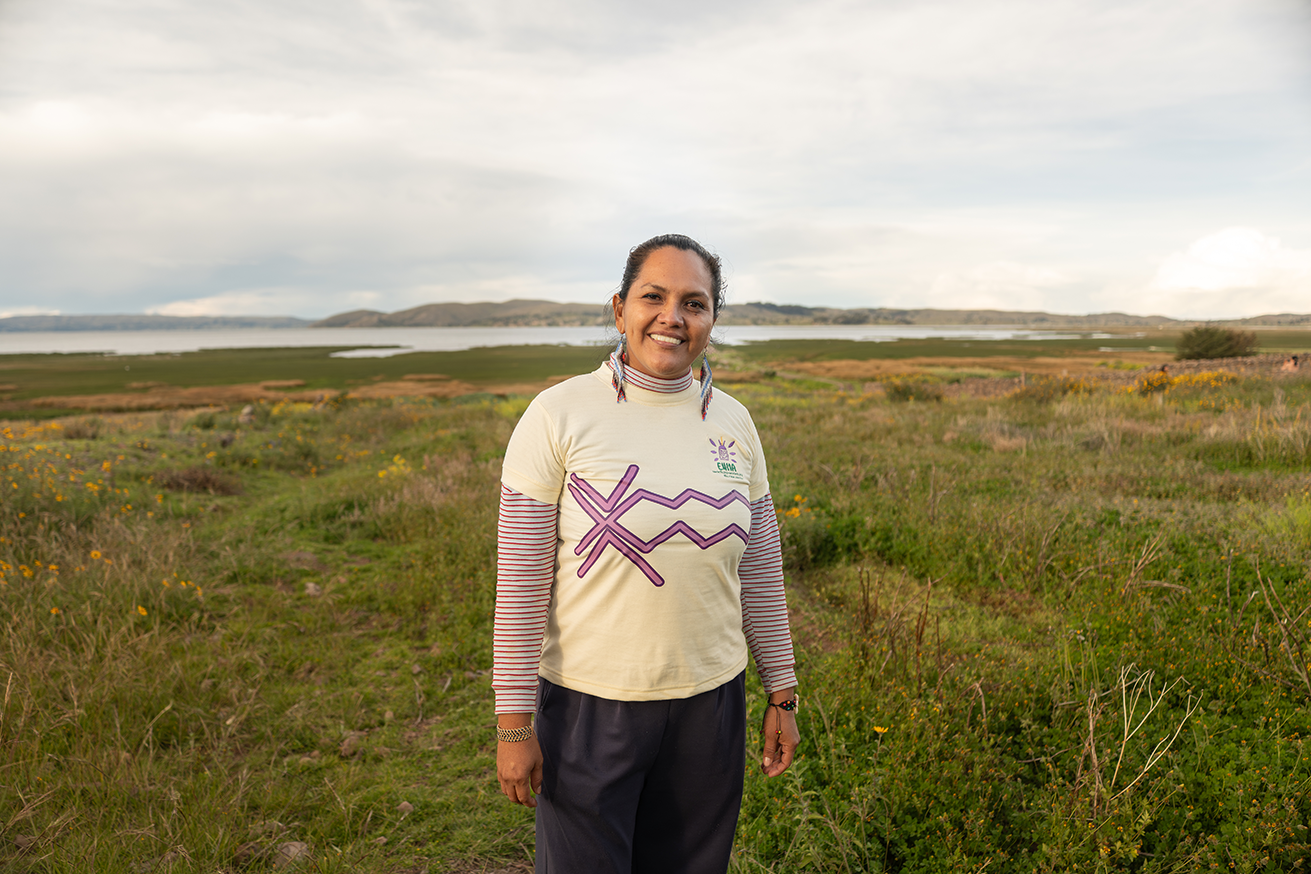 A woman stands smiling in a grassy field with a lake and hills in the background.