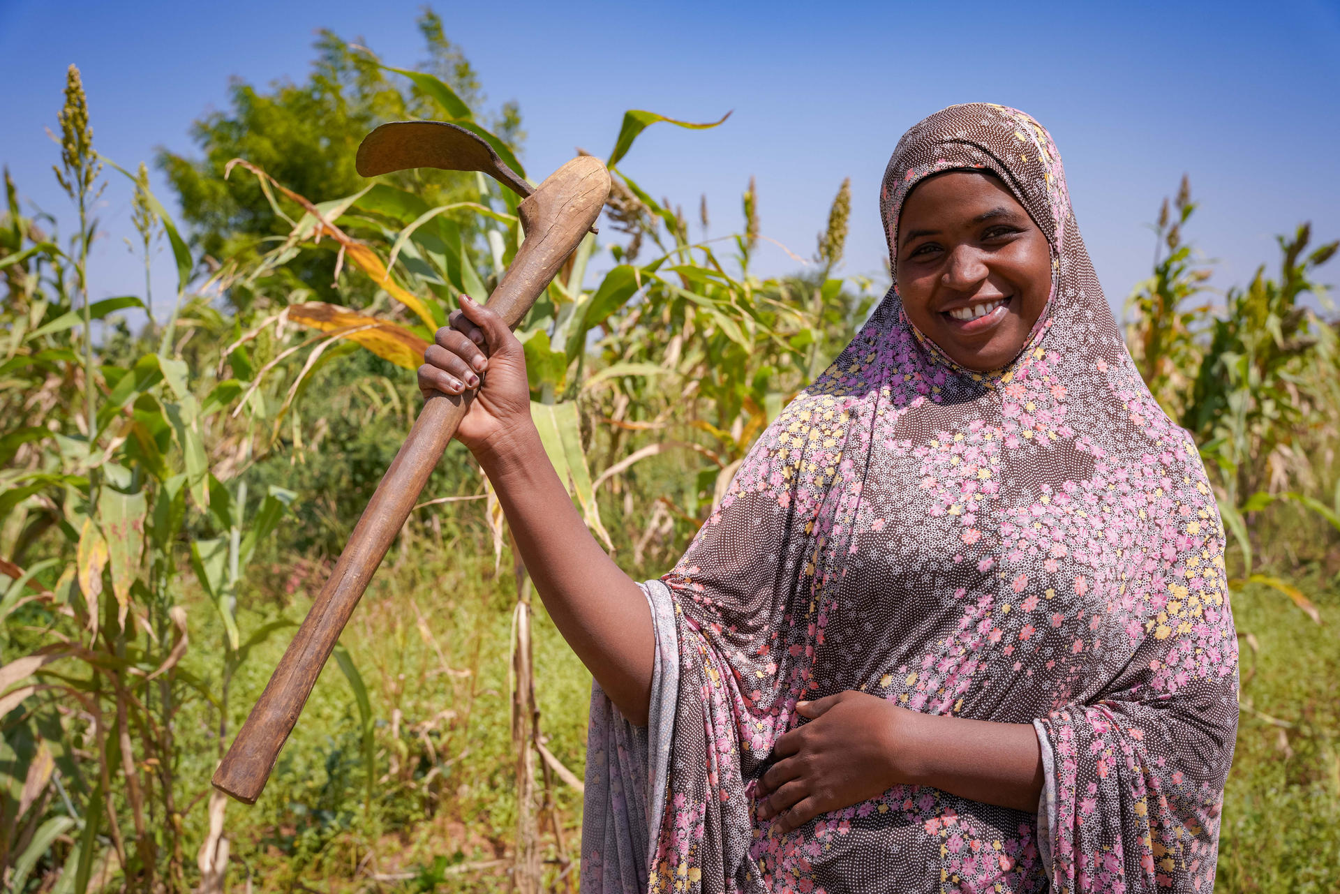 Smiling woman in a patterned hijab holds a hoe in a green field.