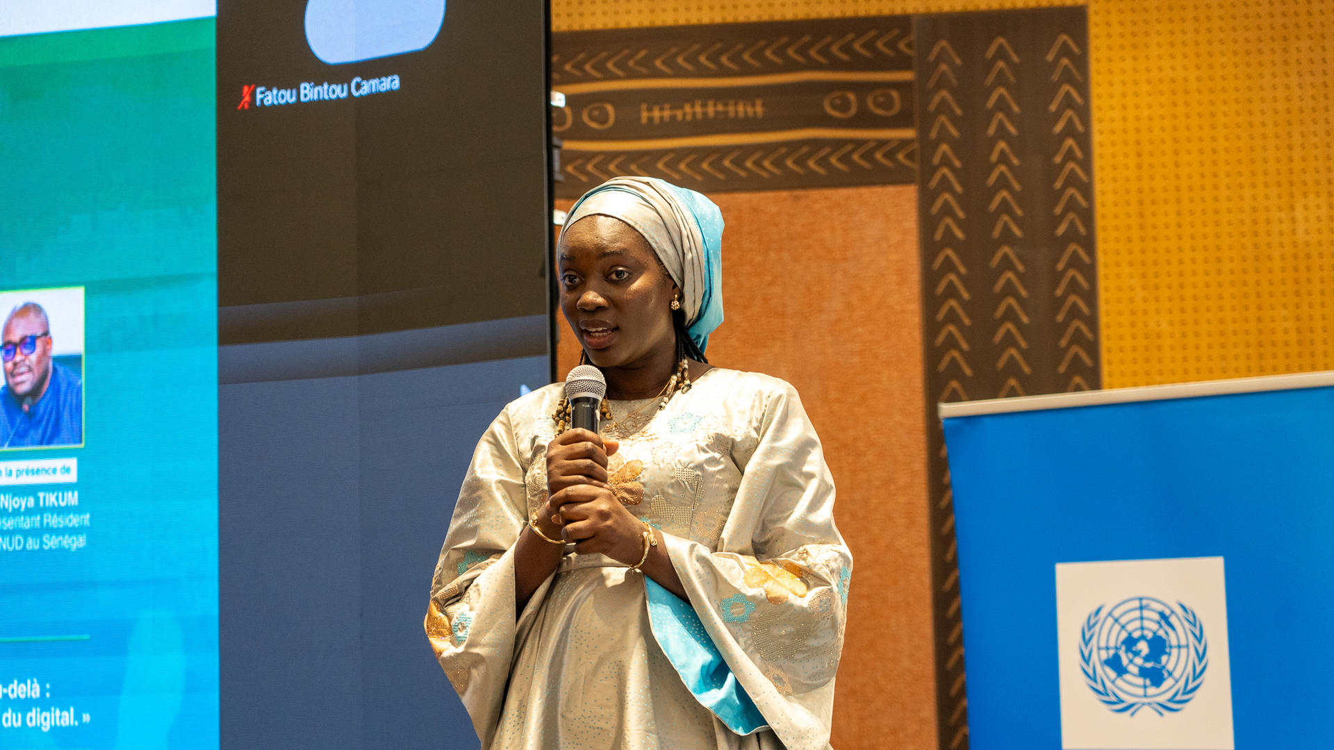 A woman in traditional attire speaks into a microphone at an event, with a digital screen behind her.
