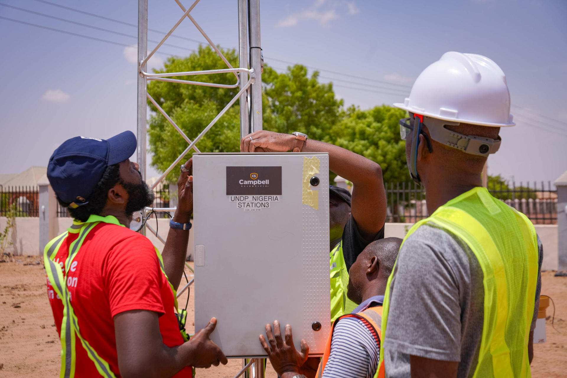 Four workers in hard hats and safety vests install equipment on a tall tower outdoors.