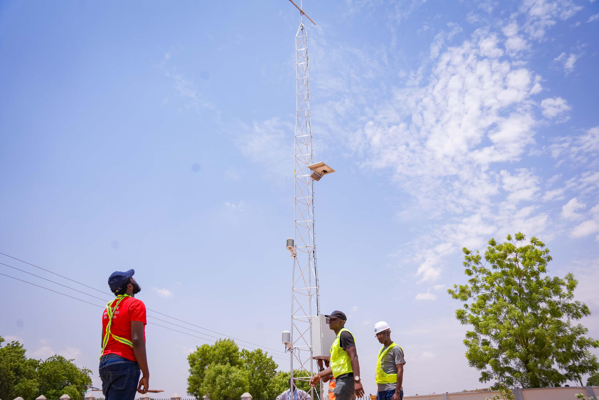 Several workers in safety gear look up at a tall tower against a blue sky.