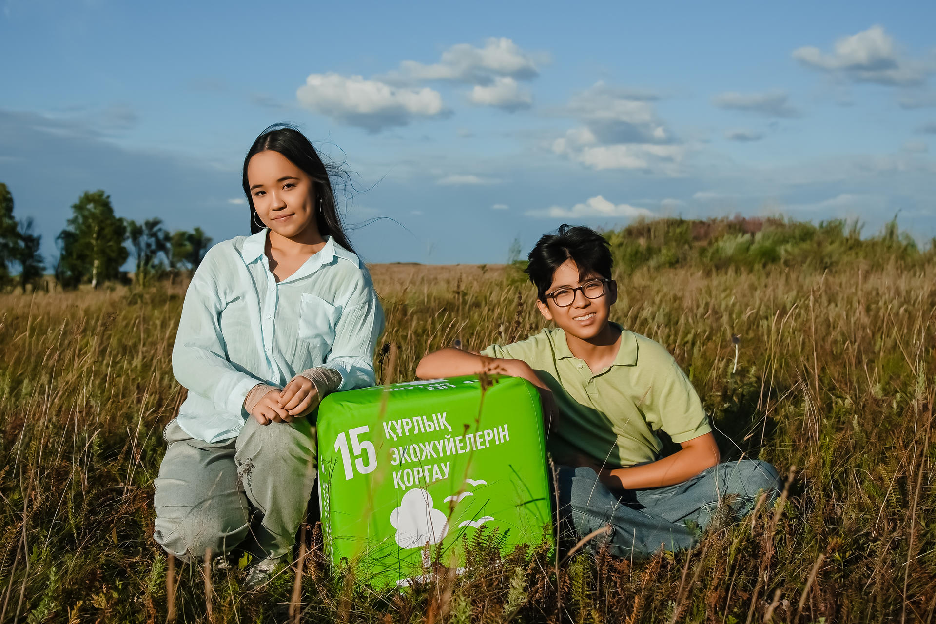 Two children pose beside a green recycling box in a grassy field under a blue sky.
