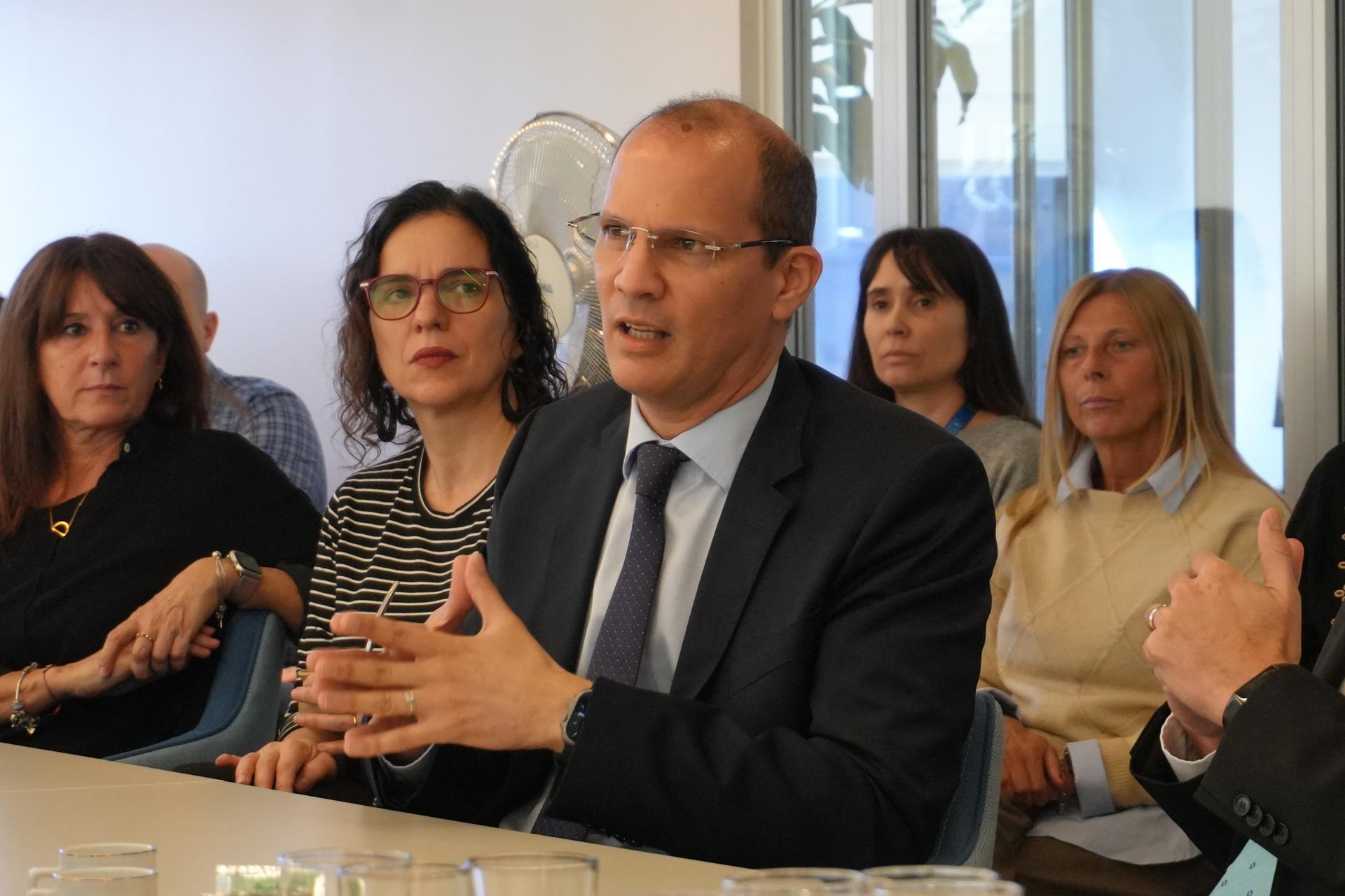 A man in a suit speaking at a table surrounded by attentive listeners.