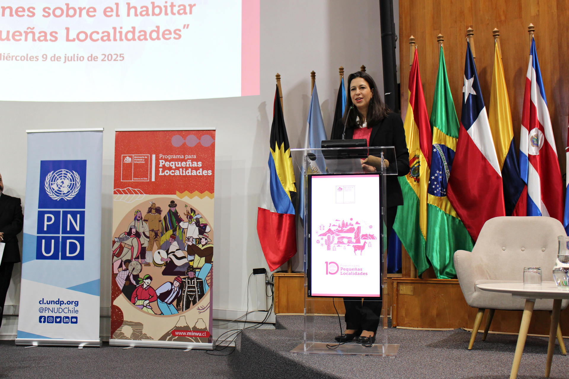 A woman speaking at a podium with flags in the background and banners on display.