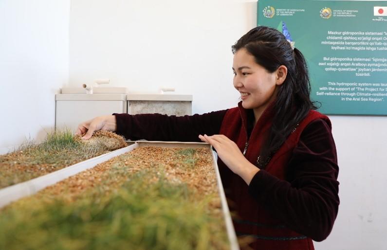 A woman in a red jacket tends to a tray of green plants, smiling in a bright indoor setting.