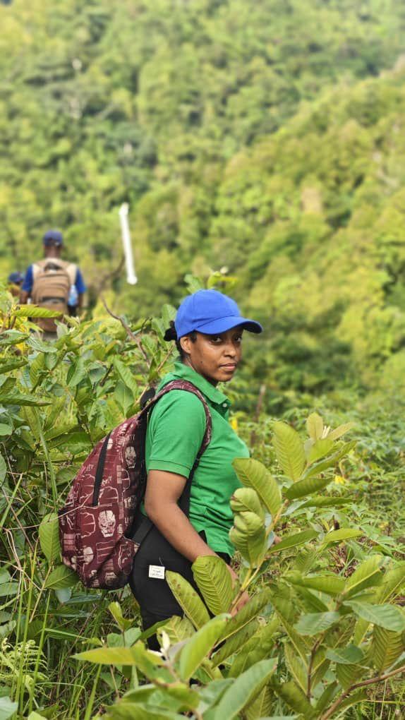 A woman in a green shirt and a blue cap smiles while hiking through lush greenery.