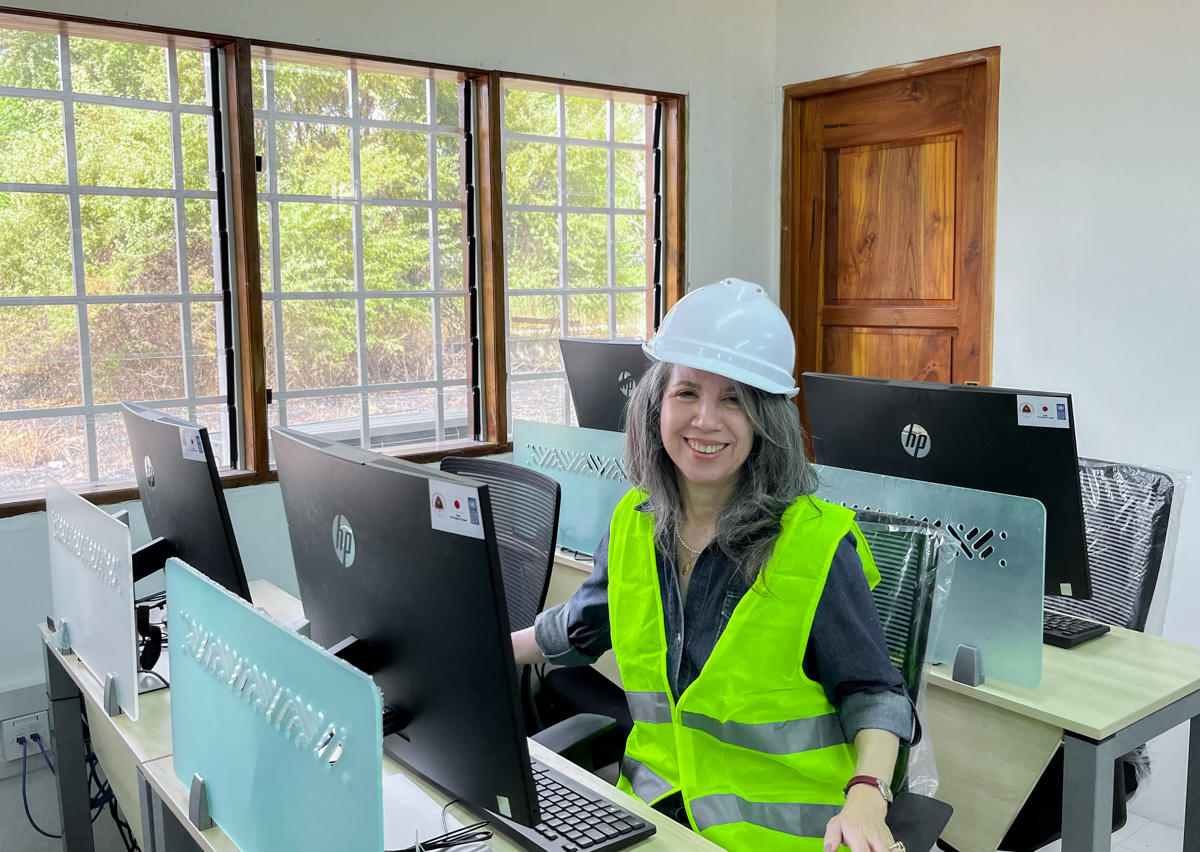 Smiling woman in a hard hat and safety vest, seated at a desk with computers.