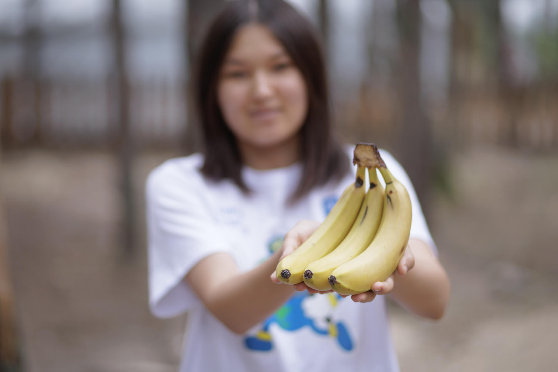 A girl in a white t-shirt holds a bunch of ripe bananas outdoors, with trees in the background.