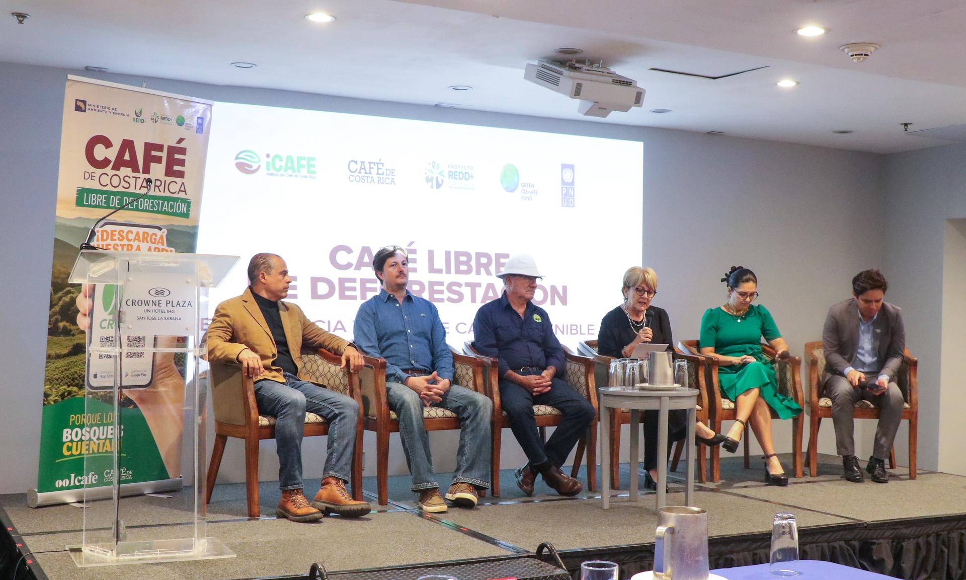 A panel of five speakers seated on a stage during an event, with a presentation backdrop.
