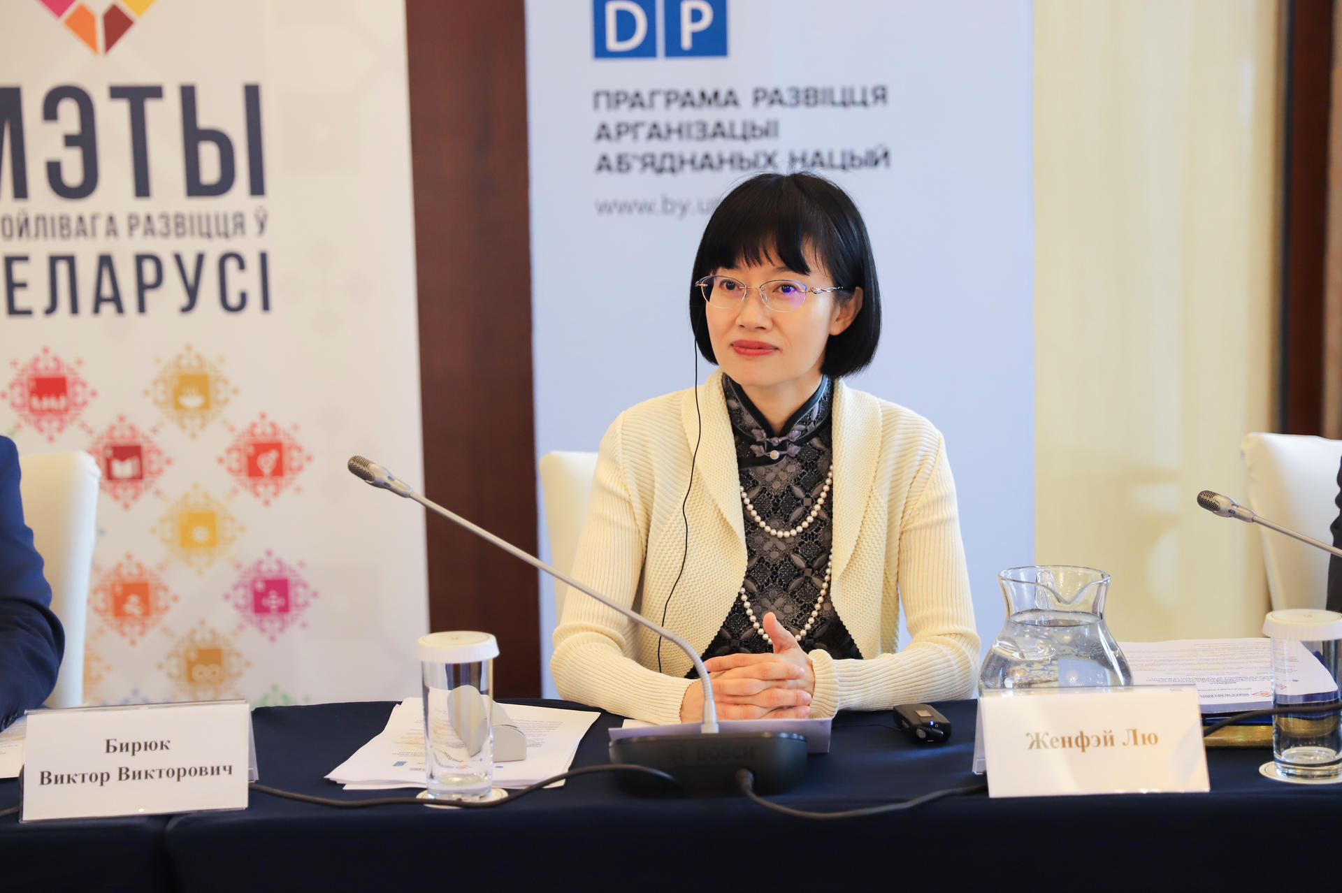 A woman with black hair and glasses sits at a conference table, looking thoughtful.