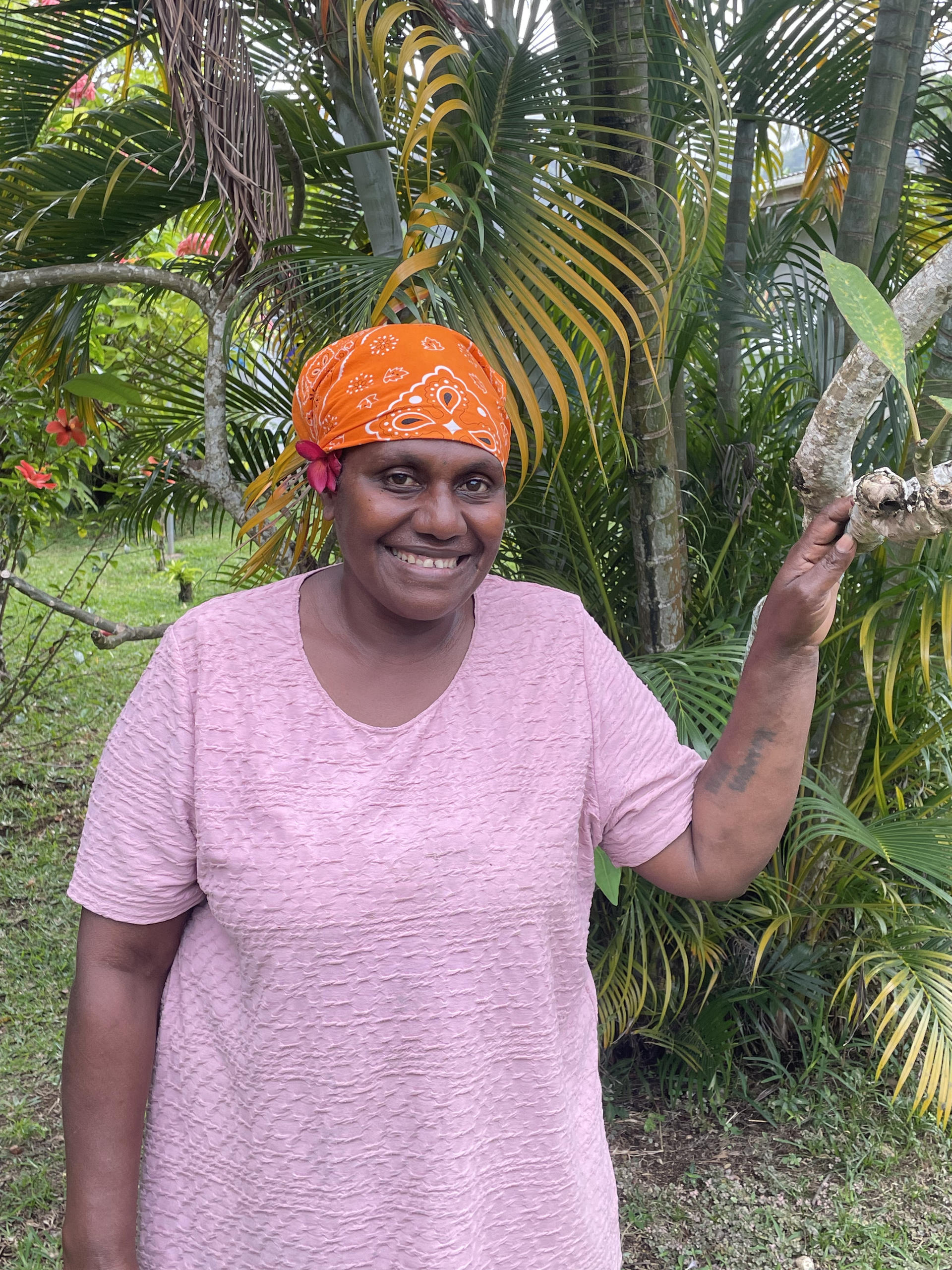Smiling woman in a pink shirt and orange headscarf standing near tropical plants.