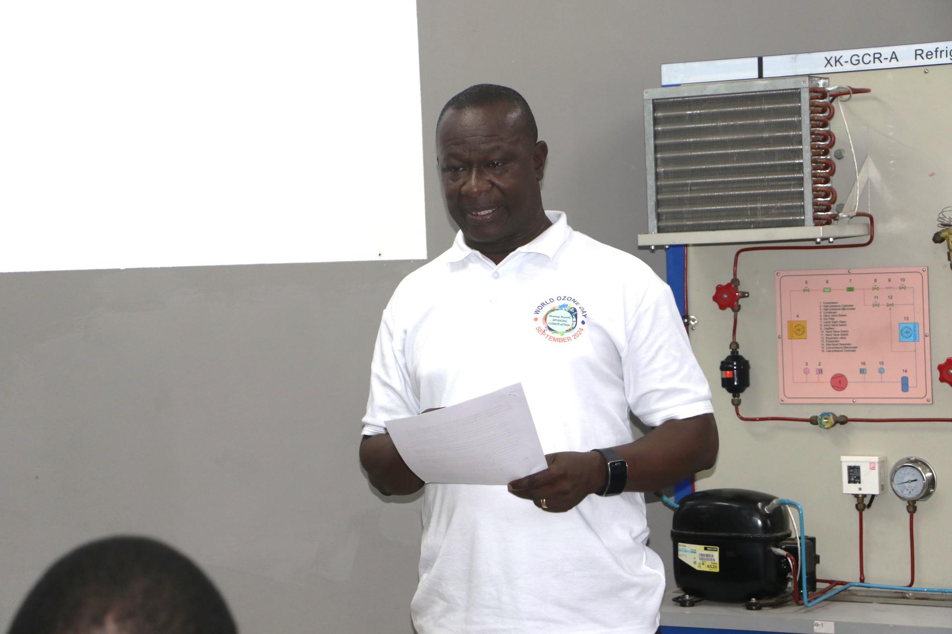 A man in a white polo shirt speaks in front of a classroom setting.