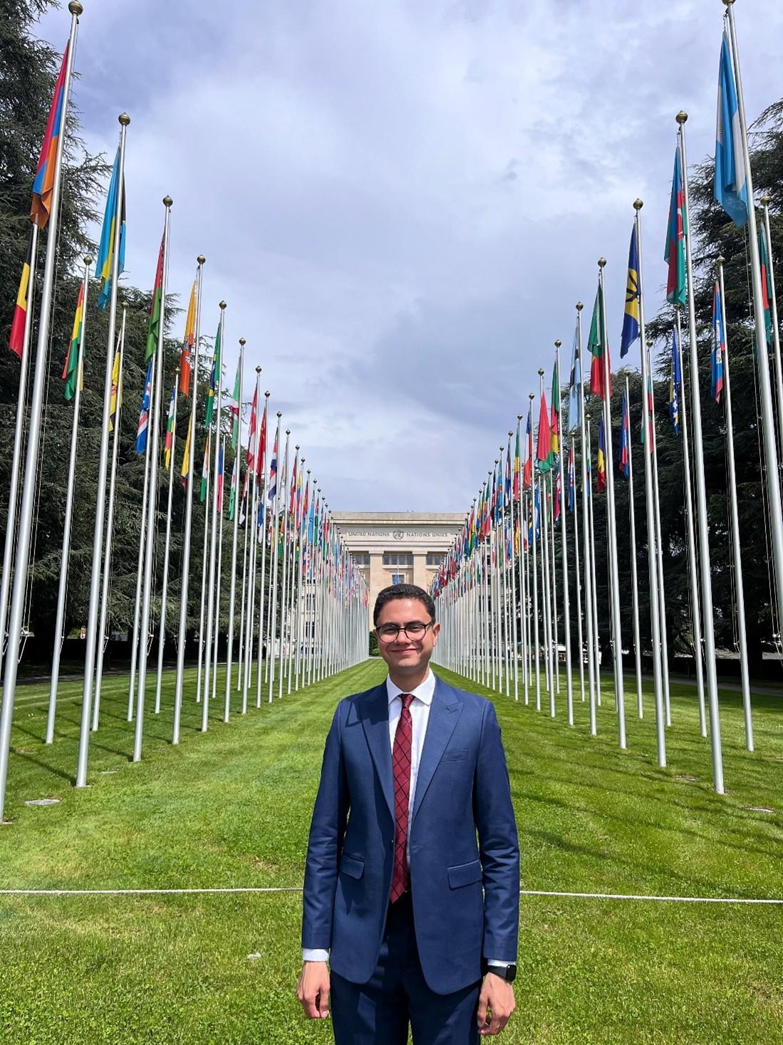 Man in a suit stands on a grassy path lined with numerous national flags under a cloudy sky.