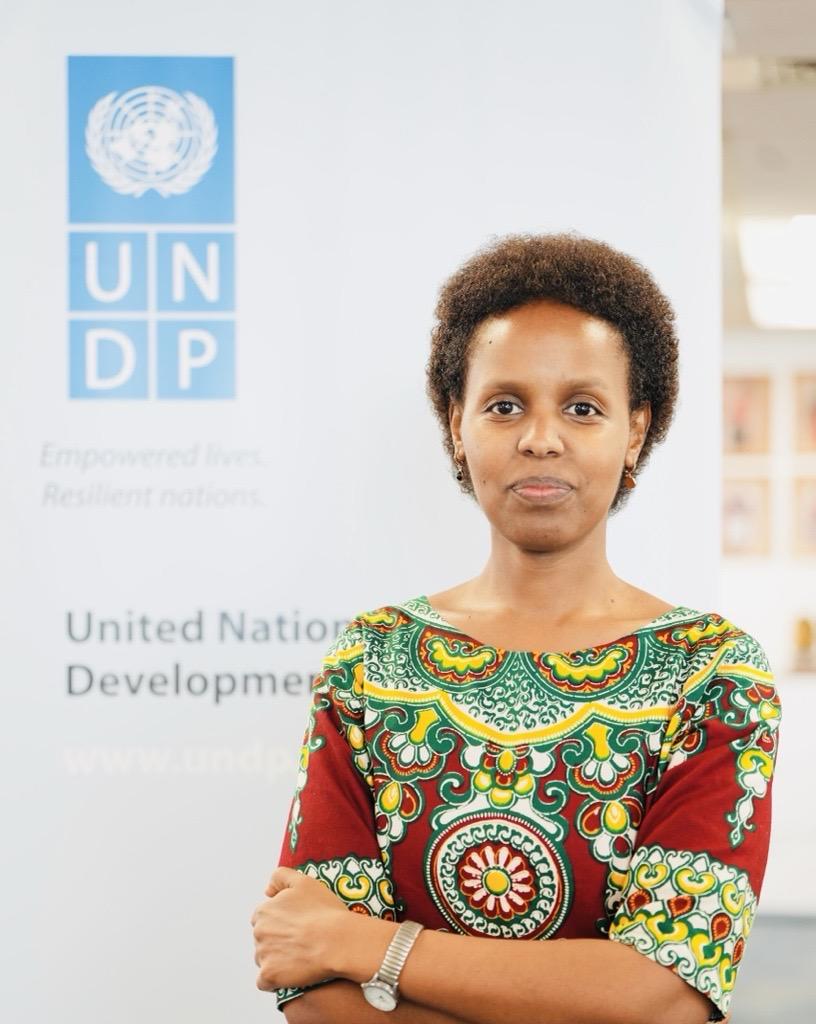 A woman in a colorful traditional dress stands confidently in front of a UNDP banner.