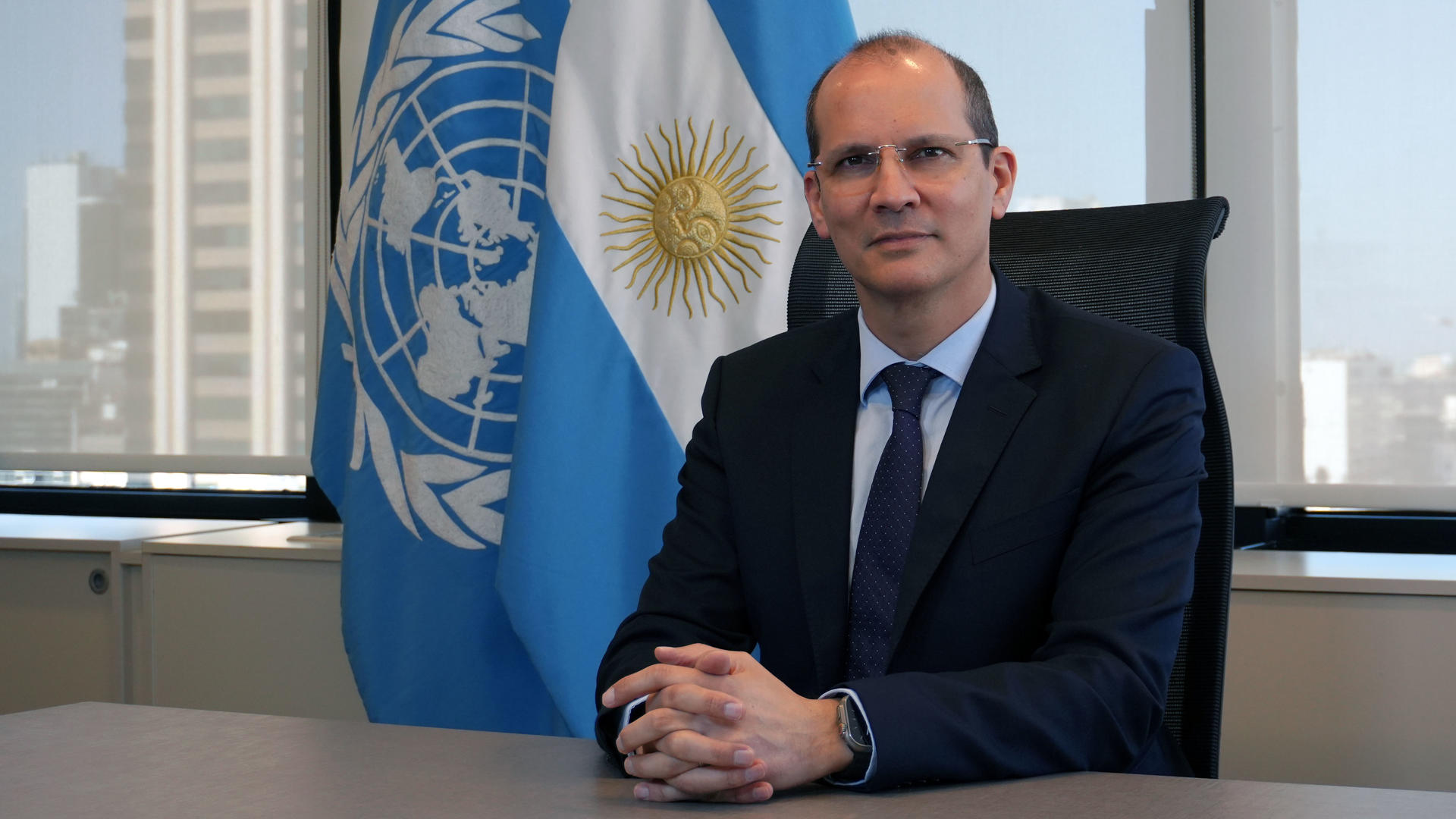 Man in a suit seated at a table, with a United Nations and Argentina flag in the background.