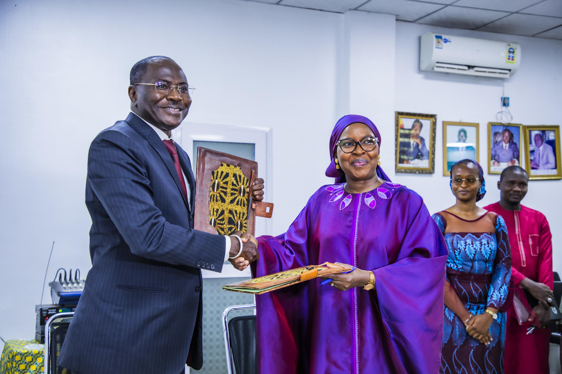 Two people shake hands while exchanging a document in a formal setting; smiles all around.
