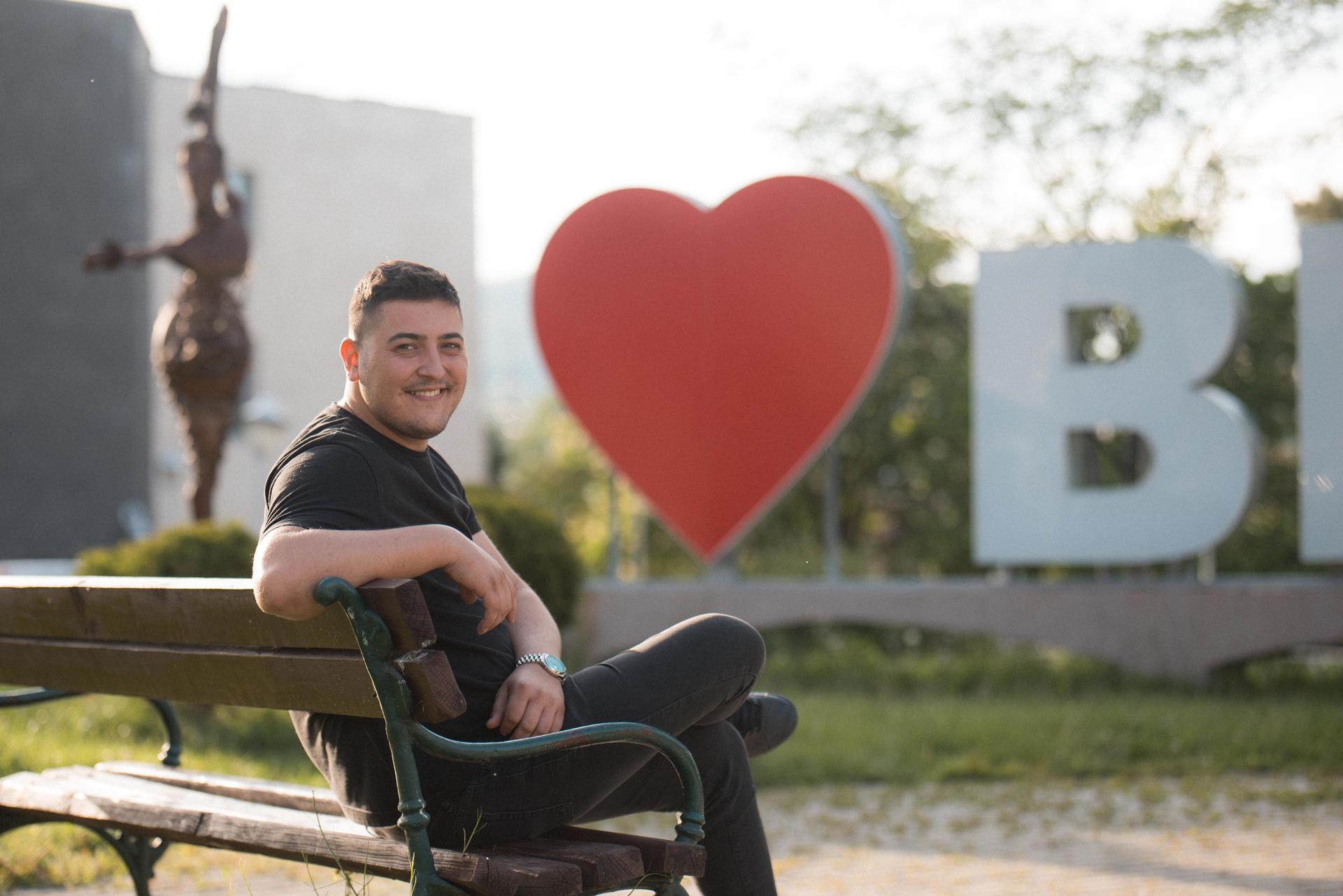 A smiling man seated on a bench with a large heart and "B" letters in the background.