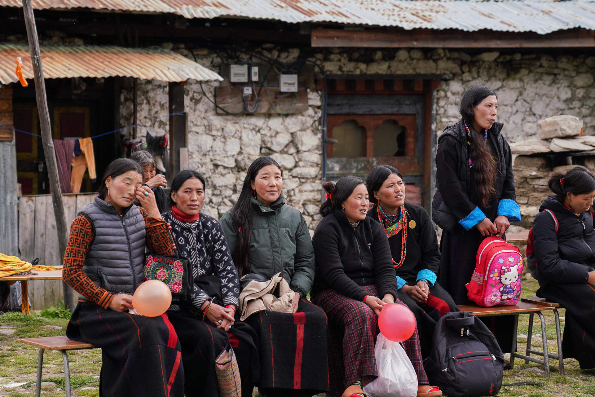 A group of women in traditional dress sitting outside a stone building, smiling and chatting.