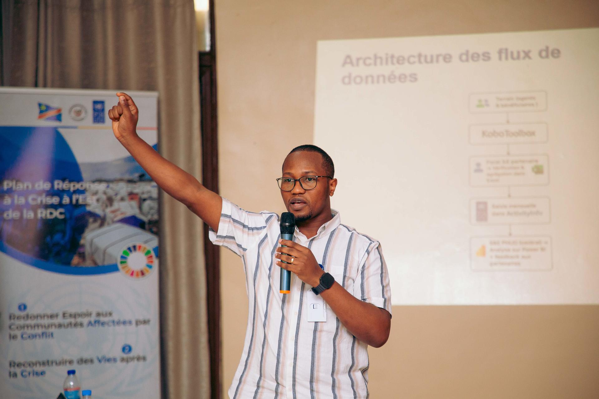 A man in a striped shirt gestures while speaking at a presentation.