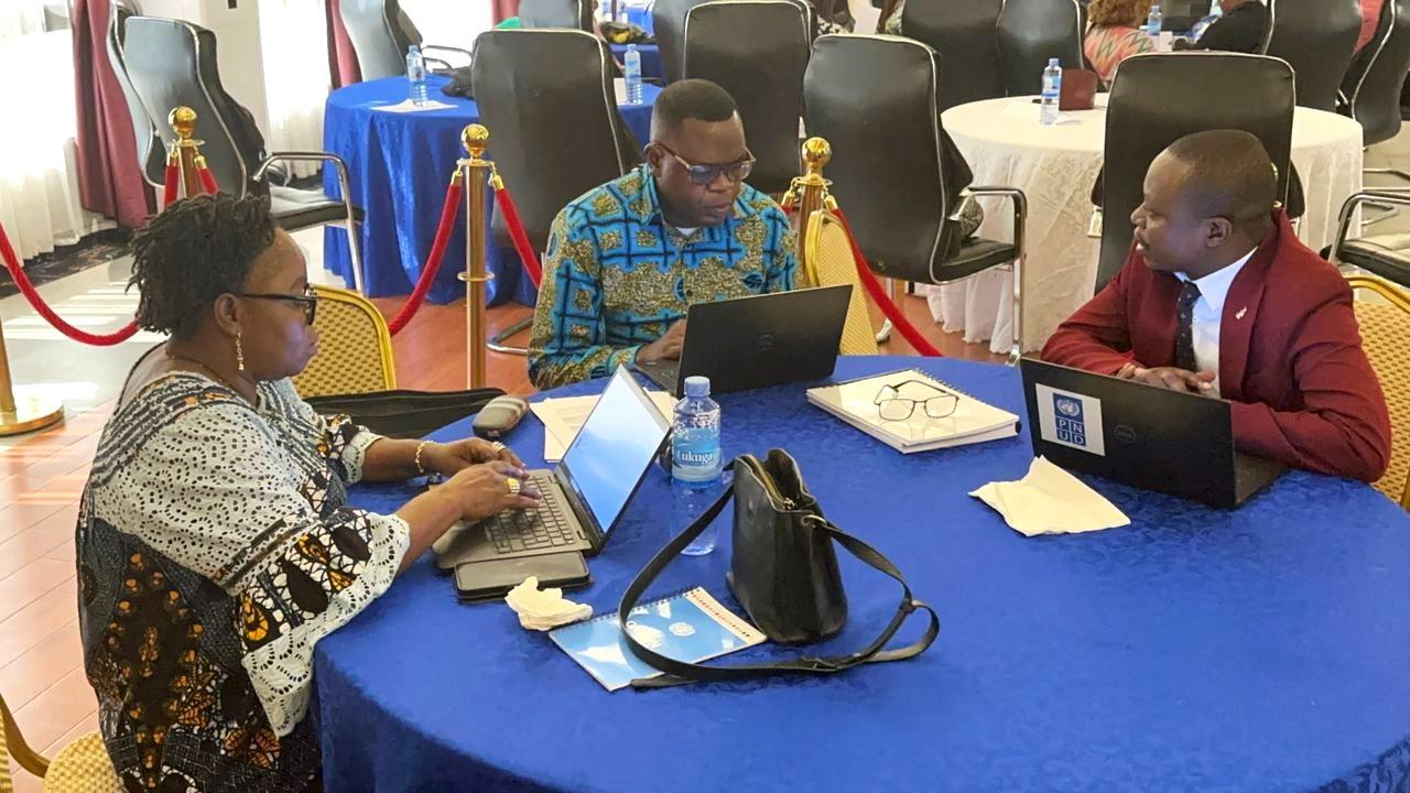 Three people seated at a blue table, working on laptops in a conference setting.