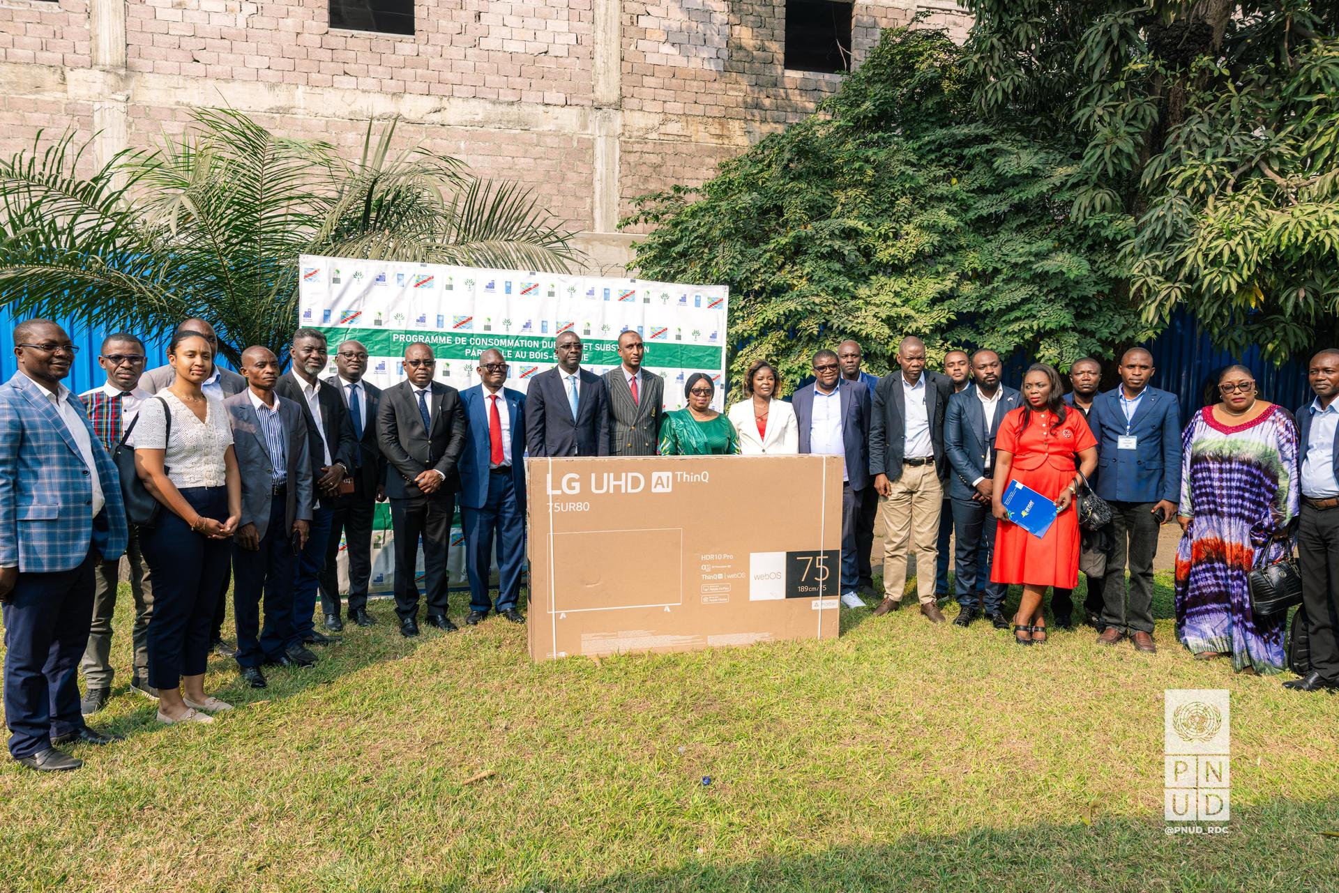 Group of people posing outdoors with a large box, likely a television, surrounded by greenery.
