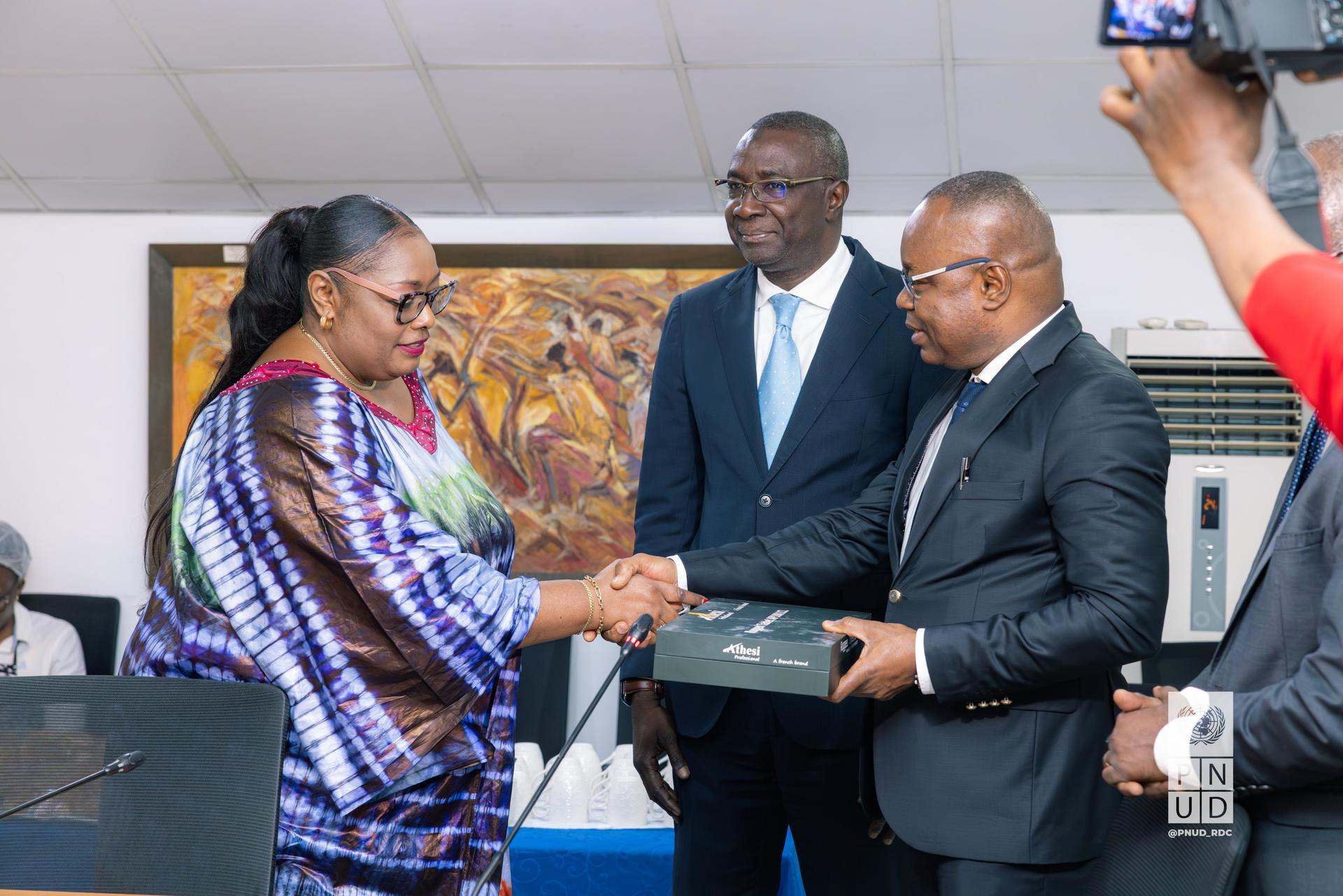 A woman in a patterned dress shakes hands with a man in a suit, exchanging documents, with two others observing.