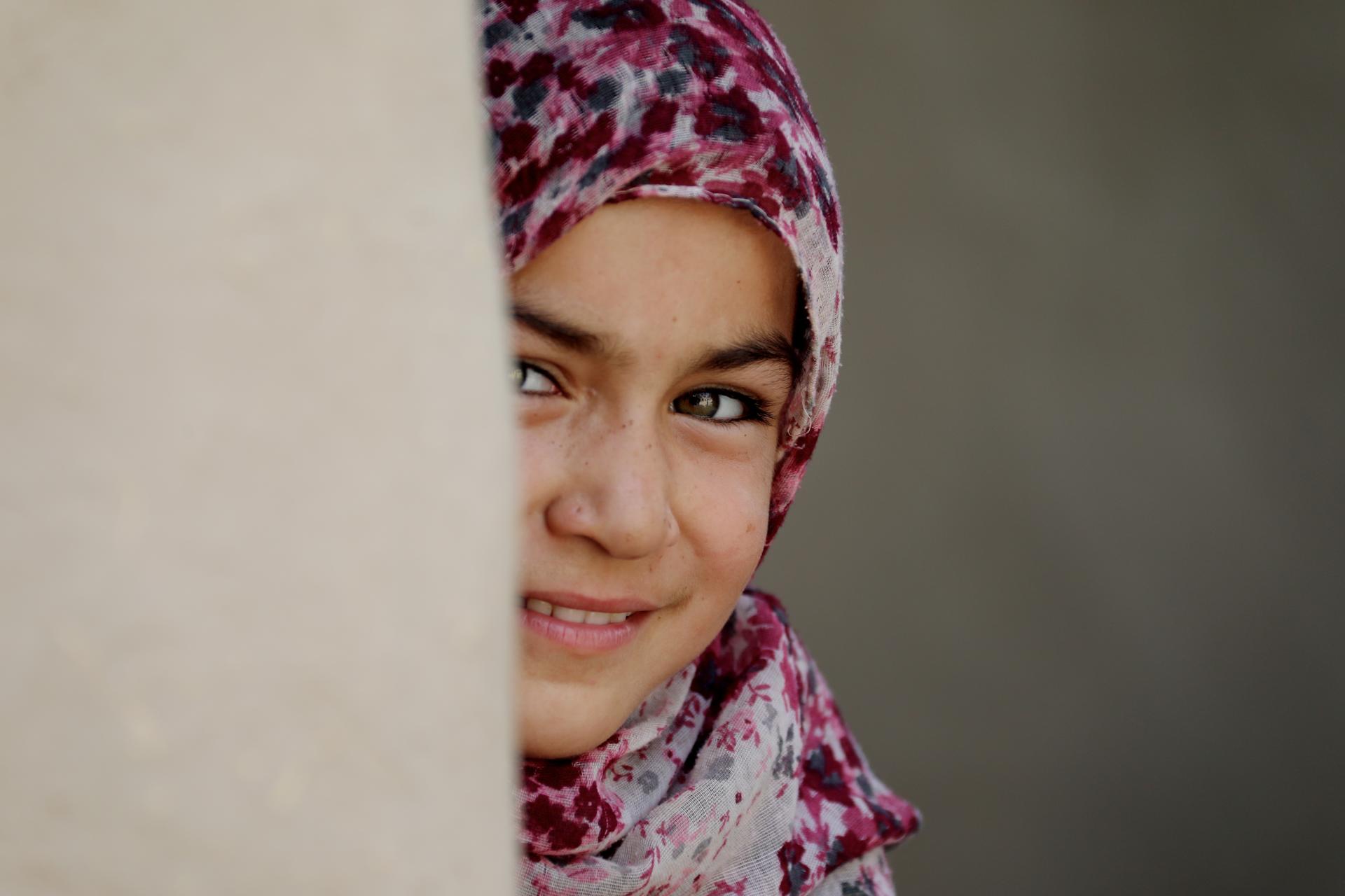 A young girl in a colorful scarf peeks out from behind a wall, smiling softly.