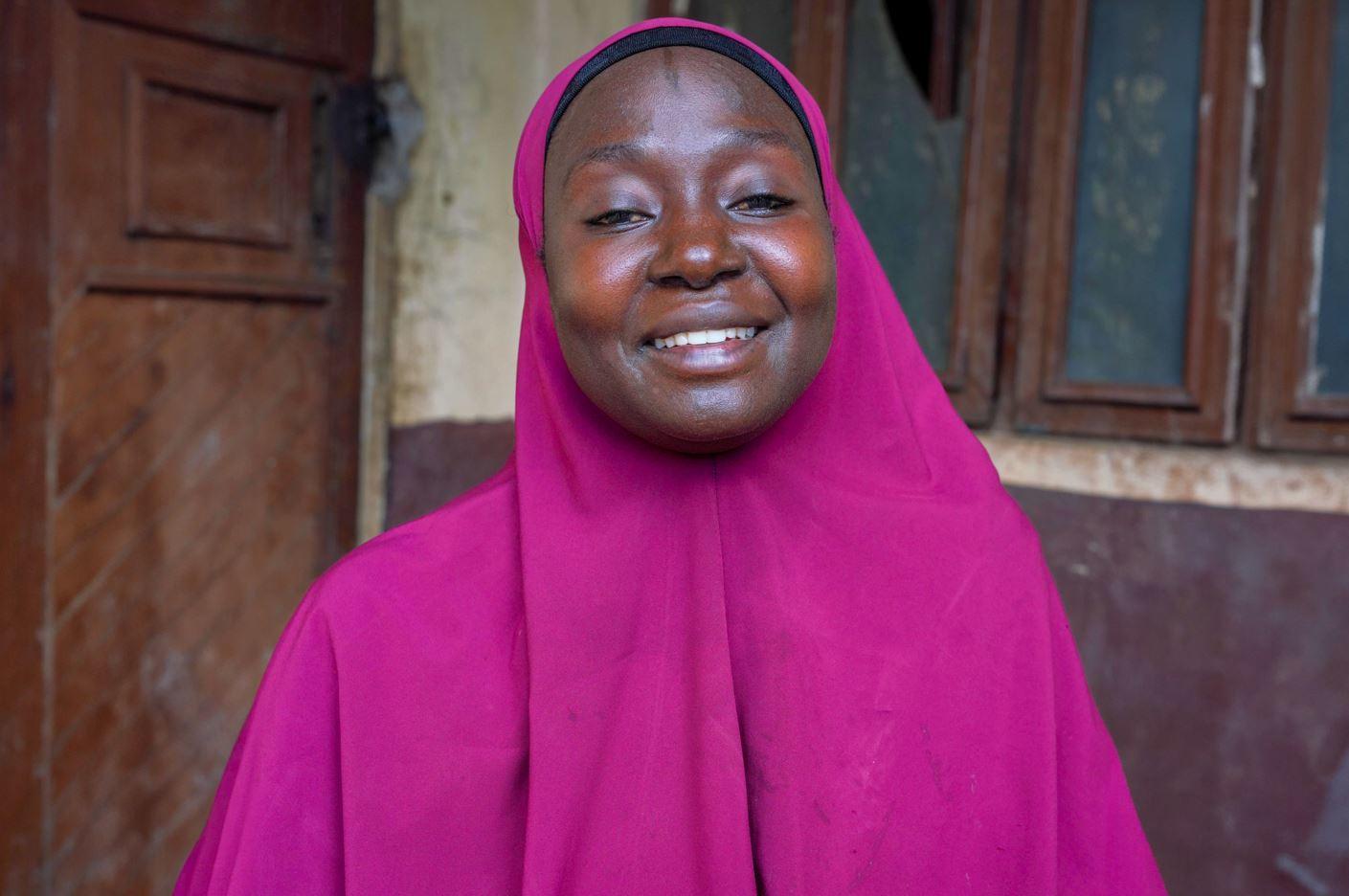 Smiling woman in a pink hijab standing outdoors near a rustic building.