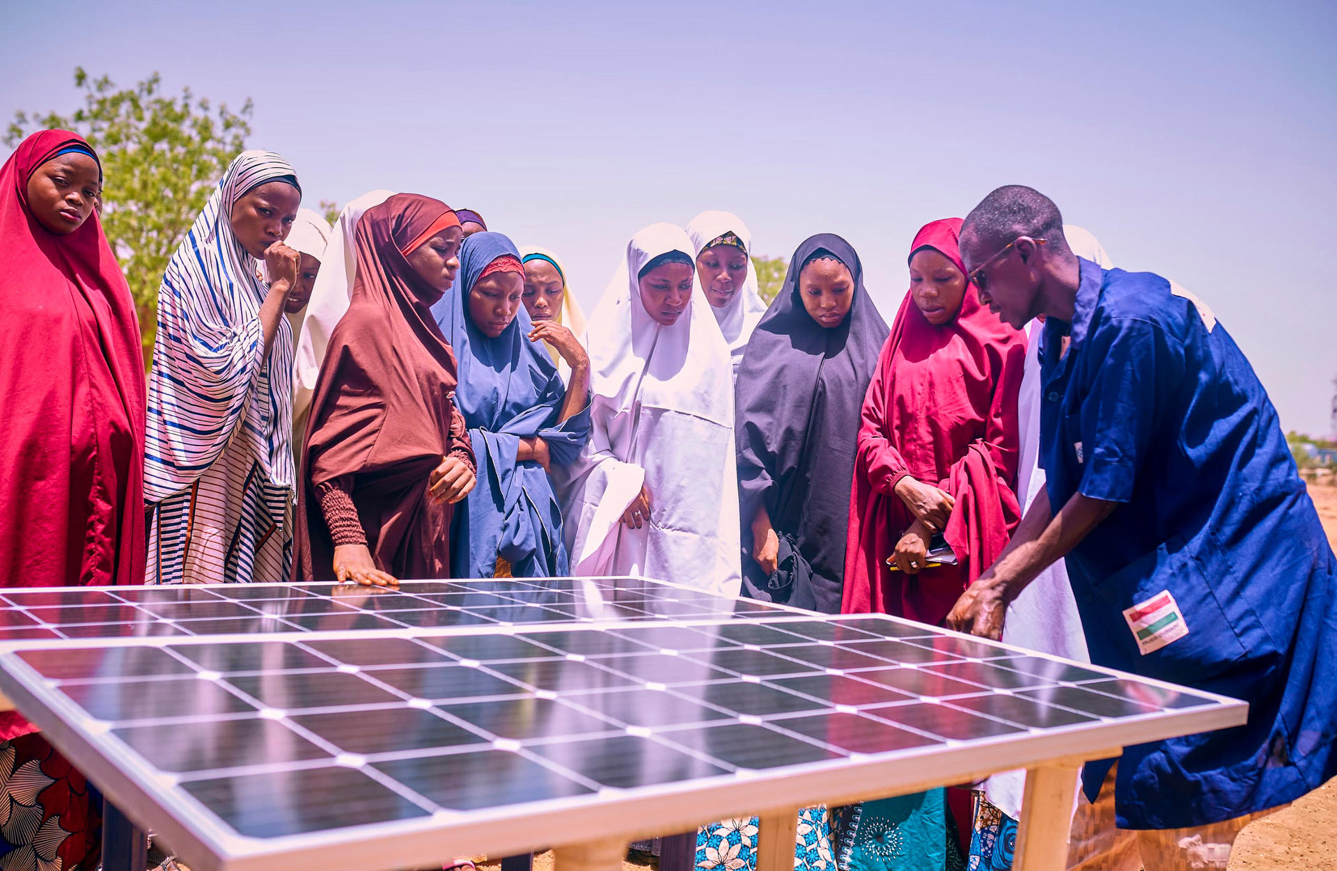 A group of women in colorful attire observes a man explaining a solar panel.