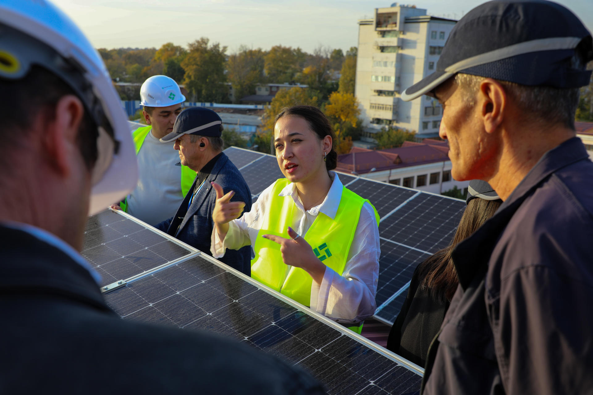 A woman in a neon vest discusses solar panels with a small group on a rooftop.