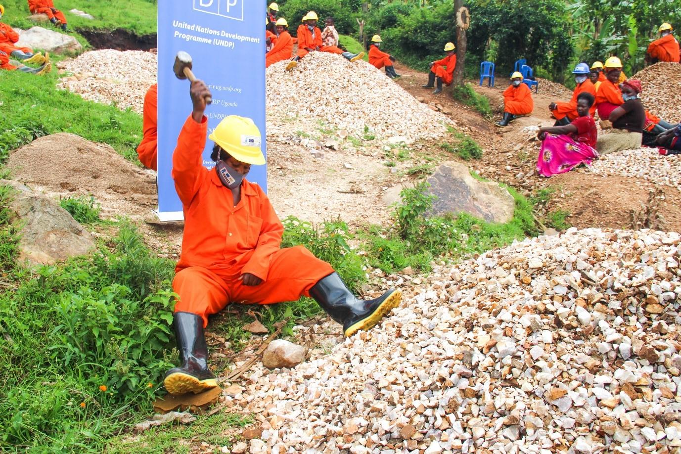 A worker in an orange jumpsuit and helmet sits on a rock pile, chipping stones, with others in the background.