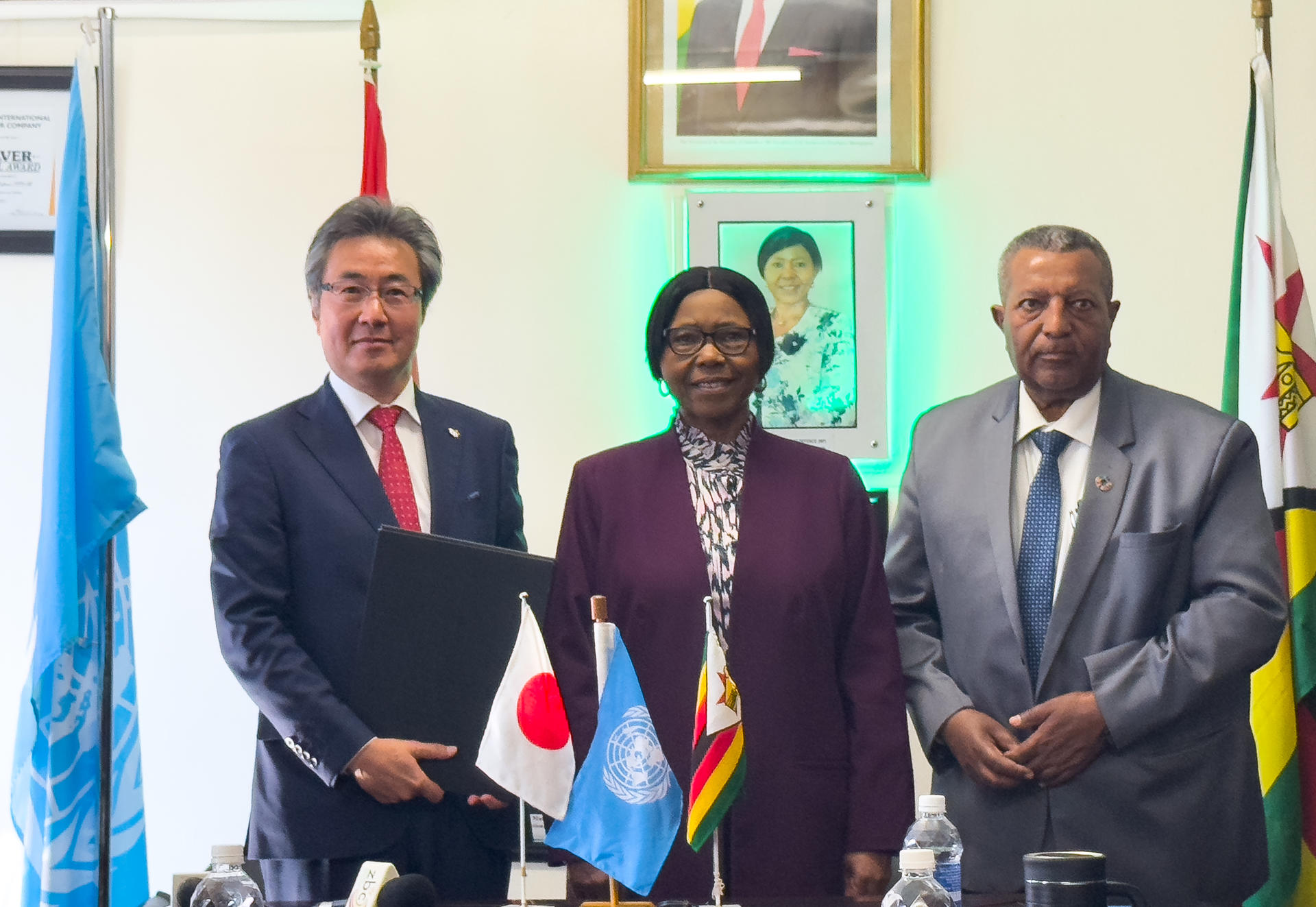 Three officials pose together at a table with flags of Japan, the UN, and Zimbabwe in the background.